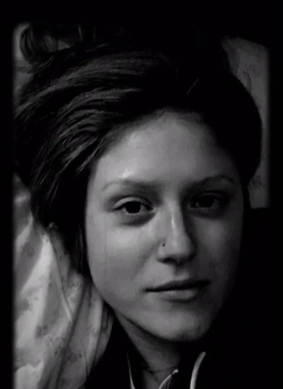 A black-and-white photo of a woman lying down, looking directly at the camera, with dark hair and a nose piercing.
