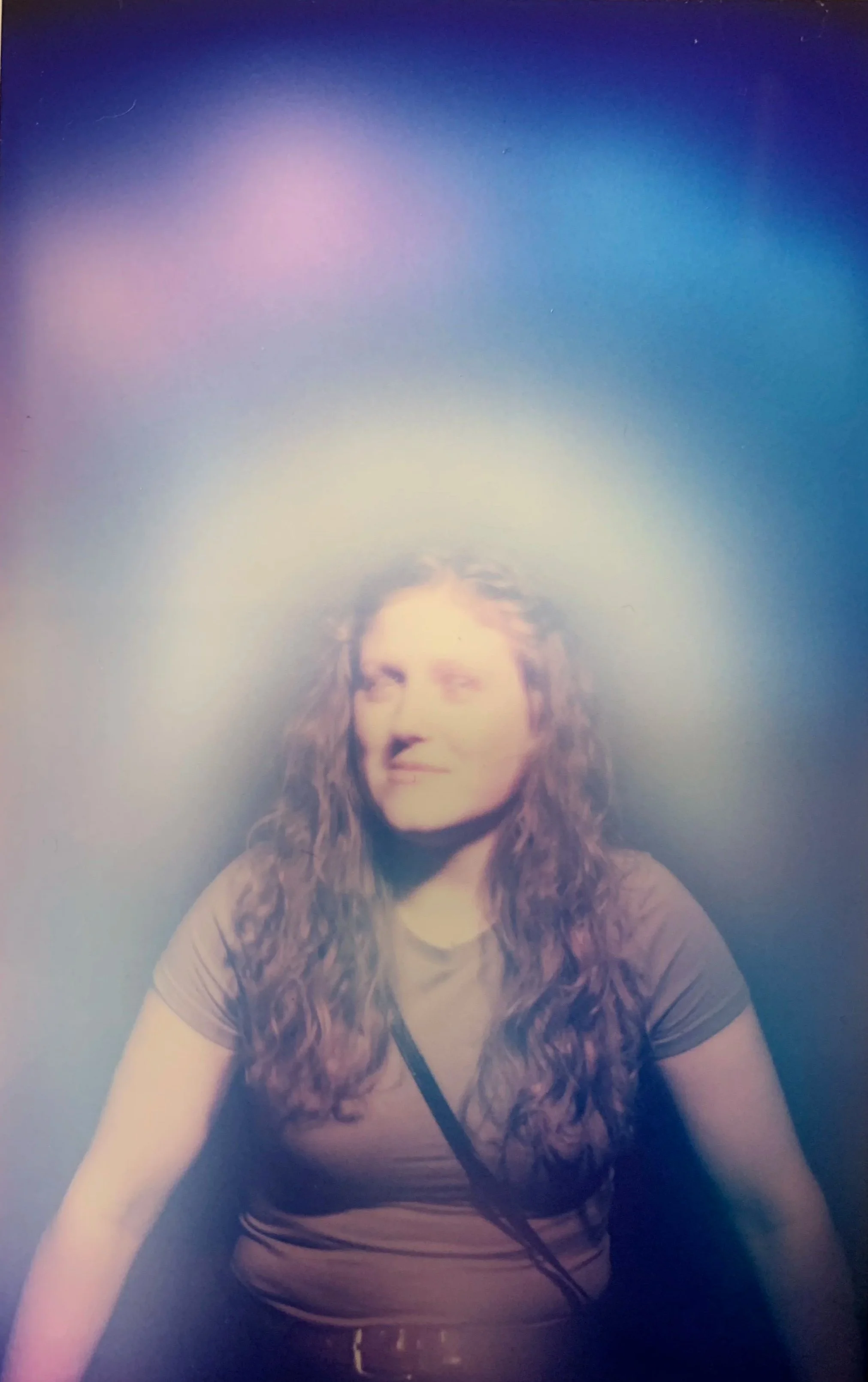 A woman with long, curly hair wearing a striped shirt. She is looking slightly to her right with a neutral expression. The background is dark with a colorful, blurred light effect above her head.