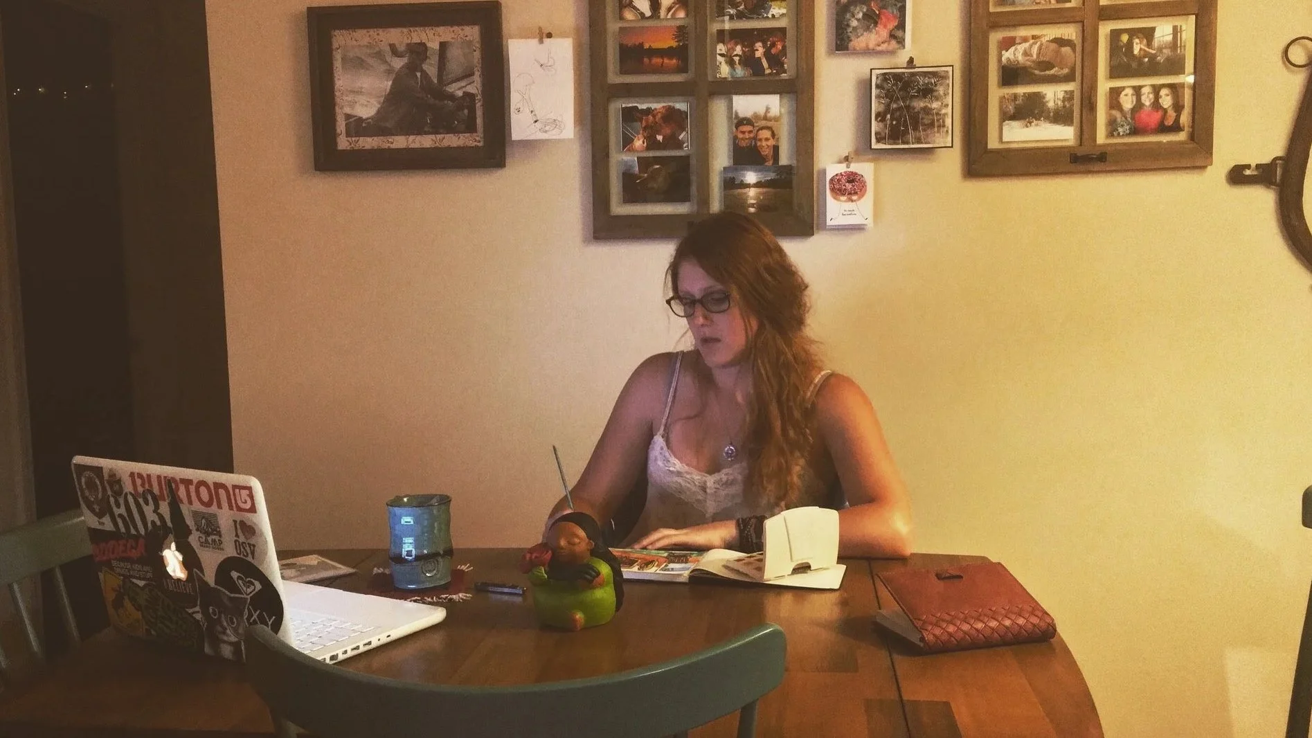 A woman with glasses and long red hair sitting at a dining table, reading a magazine or book. On the table are a laptop with stickers, a green candle holder with a figurine, a phone, a blue glass cup, and a wallet. The wall in the background has several framed photos and artwork.