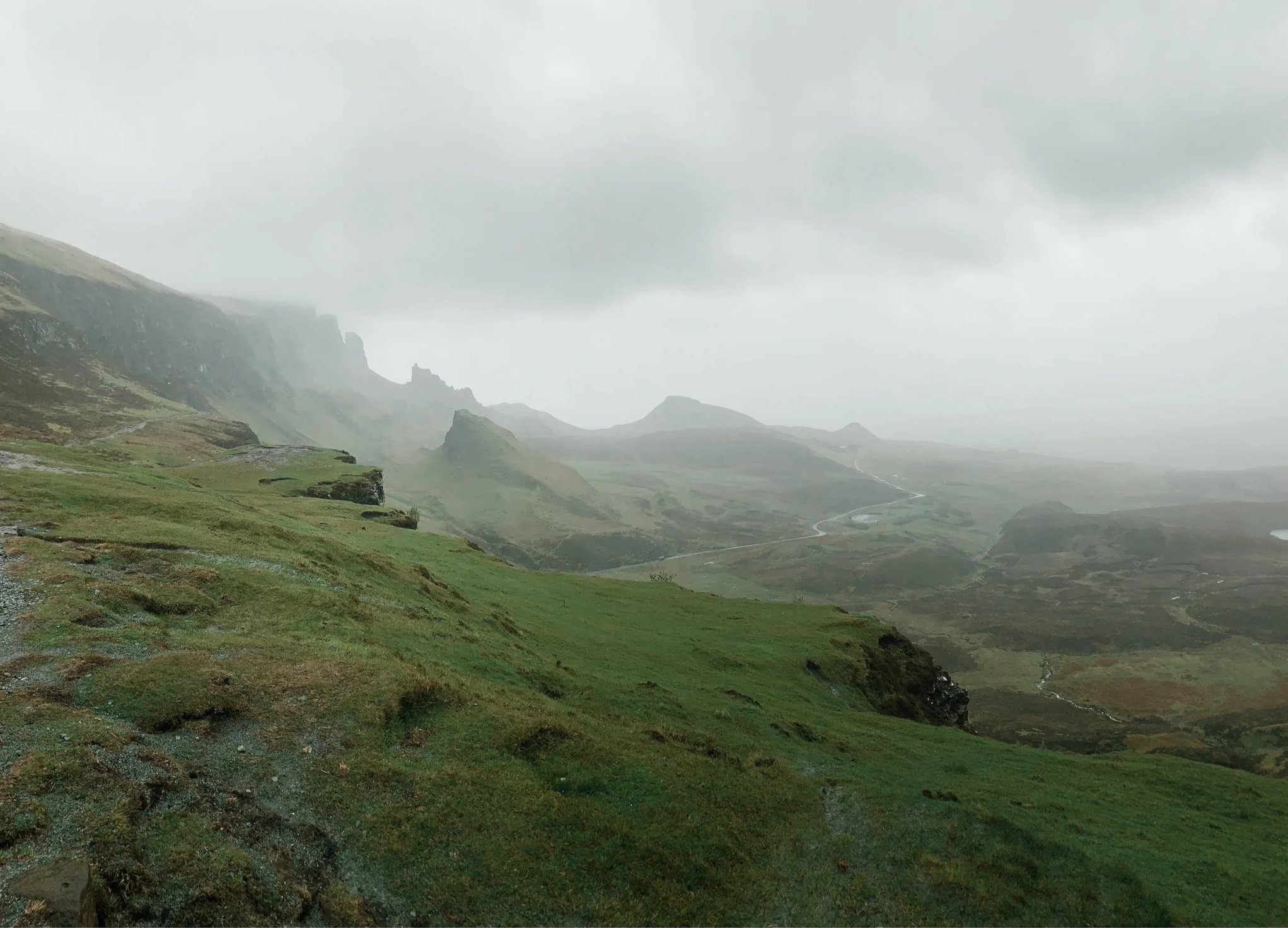 Isle of Skye. A foggy mountainous landscape with green meadow in the foreground, rocky peaks in the background, and a winding road through the valley.