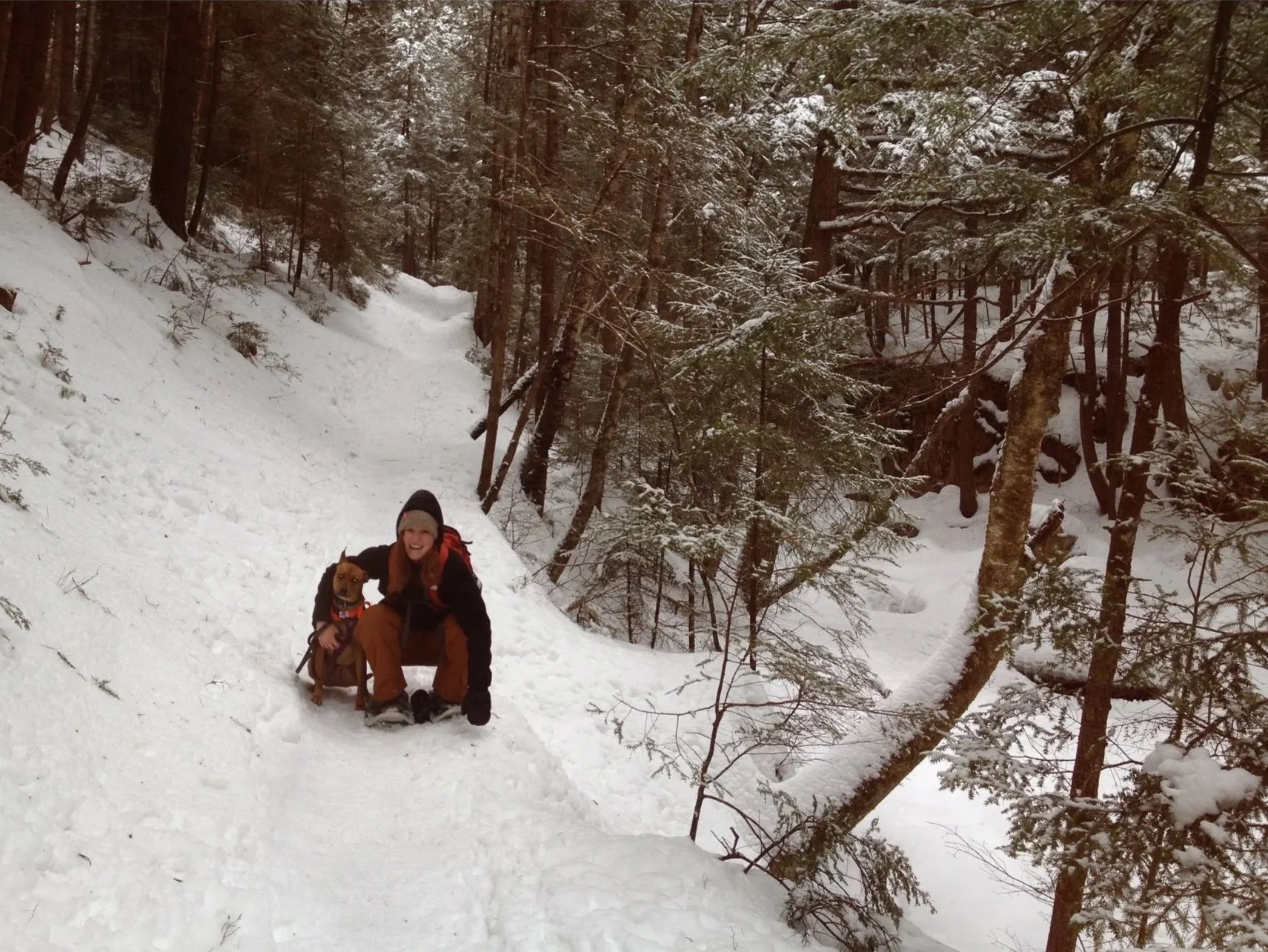 A person in winter clothing smiling while kneeling down and holding a dog in a snow-covered forested area.