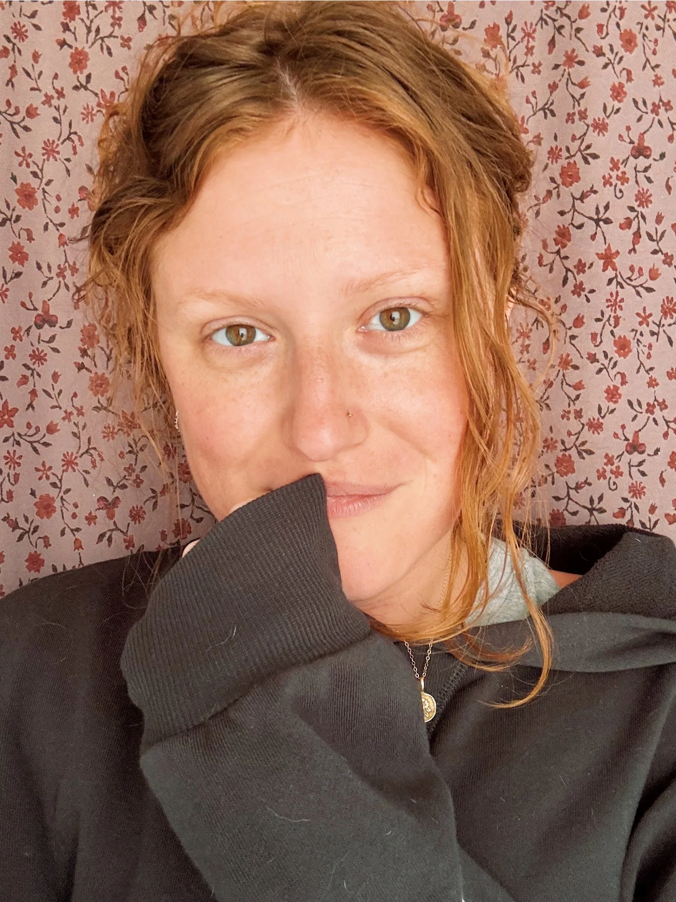 A woman with light skin, red hair, green eyes, a nose piercing, and a necklace. She is partially covering her face with her sleeve, looking at the camera, against a background of red and pink floral wallpaper.