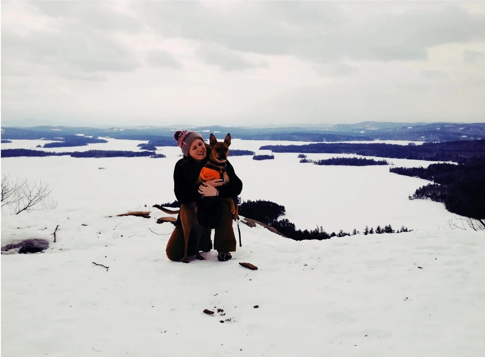 A woman in winter clothing holding a dog on a snowy mountain top overlooking a snow-covered lake or river with forested hills in the background.