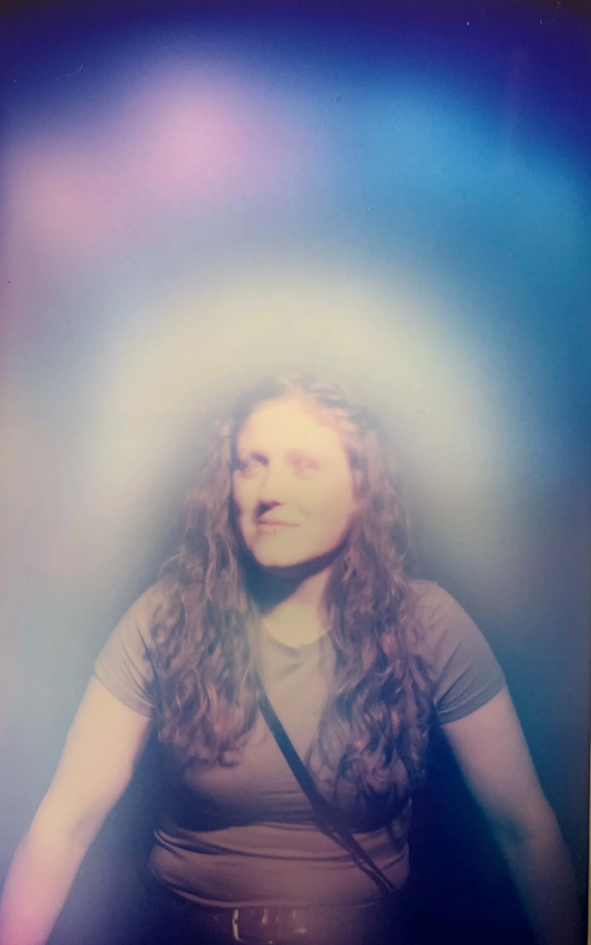 Aurora image- white aurora in & around her- indigo colored up above- pink and blue in the upper ribbon. A woman with long curly hair is smiling and looking slightly to the side. She is wearing a brown shirt and has a black strap across her shoulder.
