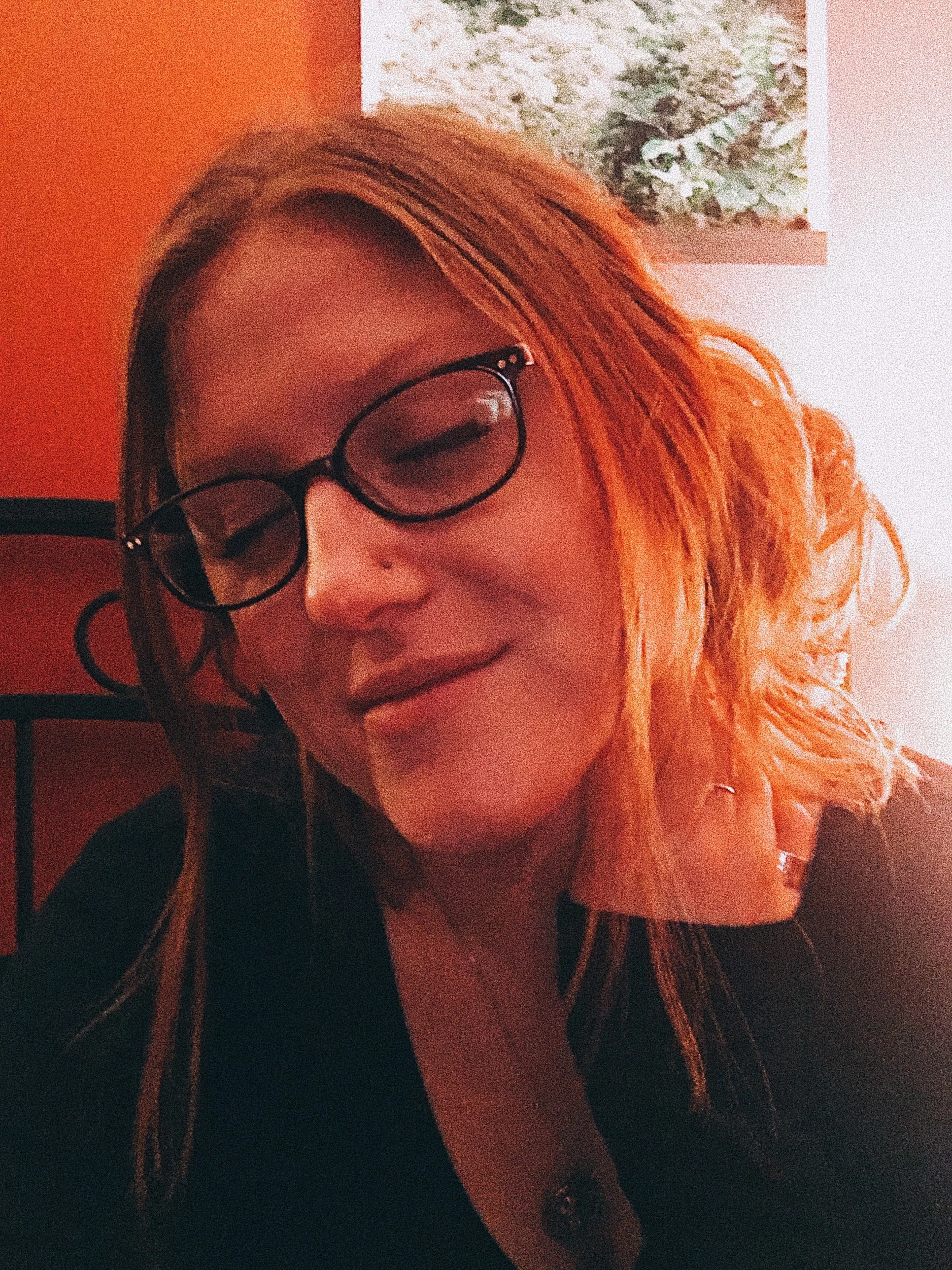 A woman with long hair and glasses smiling, next to a wall with a piece of art, in a cozy indoor setting.