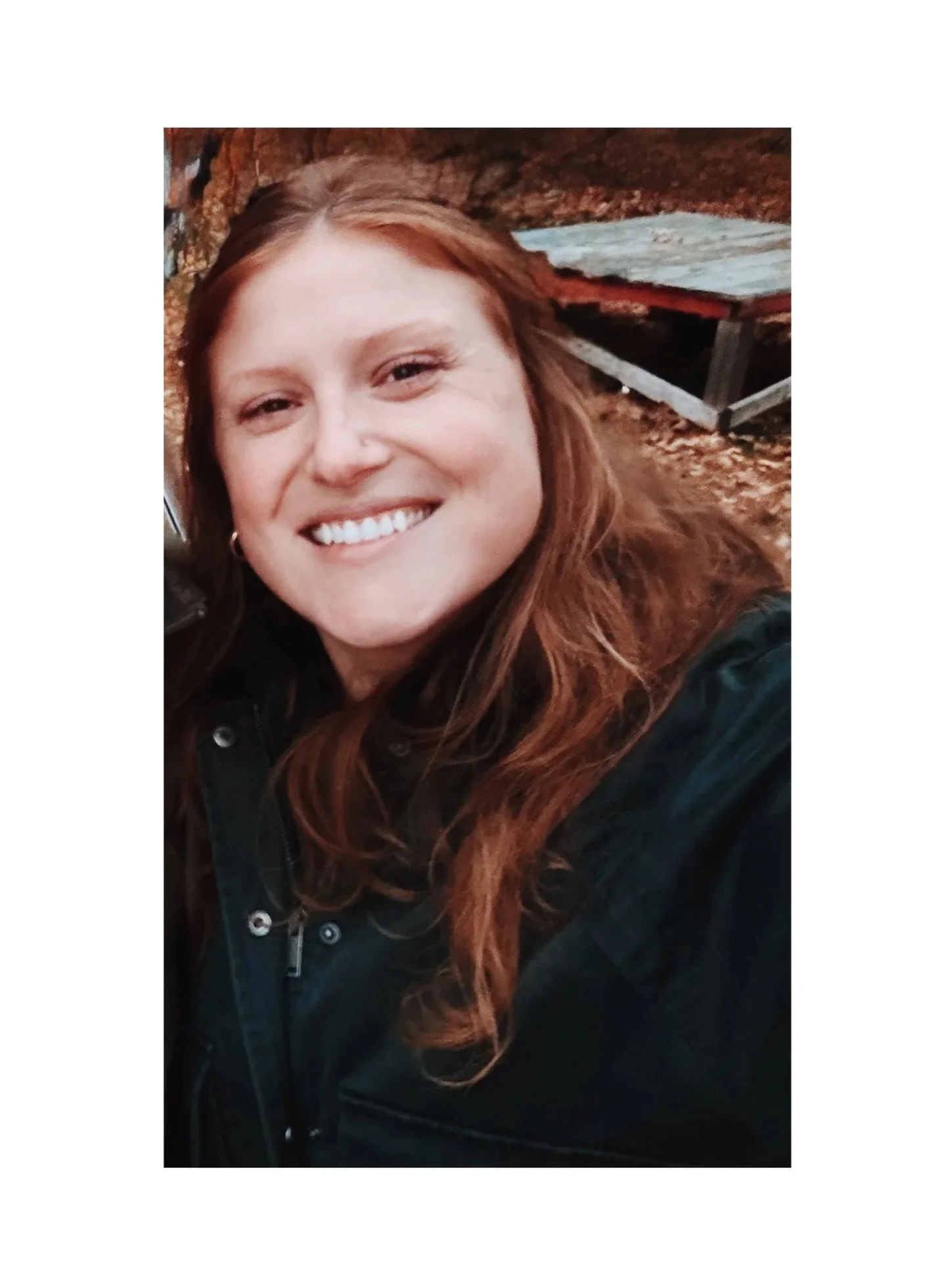 A smiling woman with long red hair, wearing a black jacket, outdoors with a leaf-covered ground and a small wooden structure in the background.