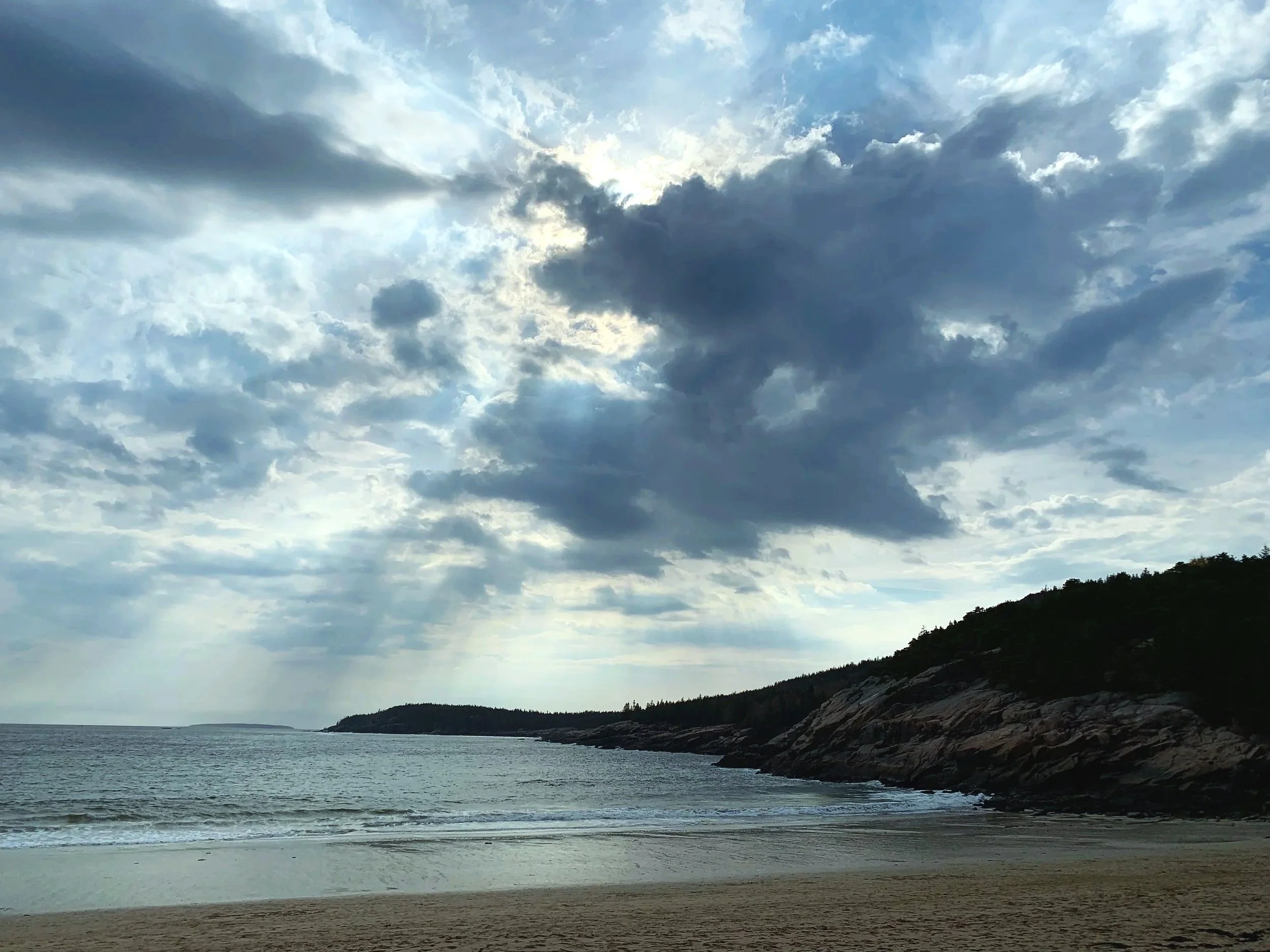 Cloudy sky over a shoreline with rocky terrain and water, with distant land and trees in the background.