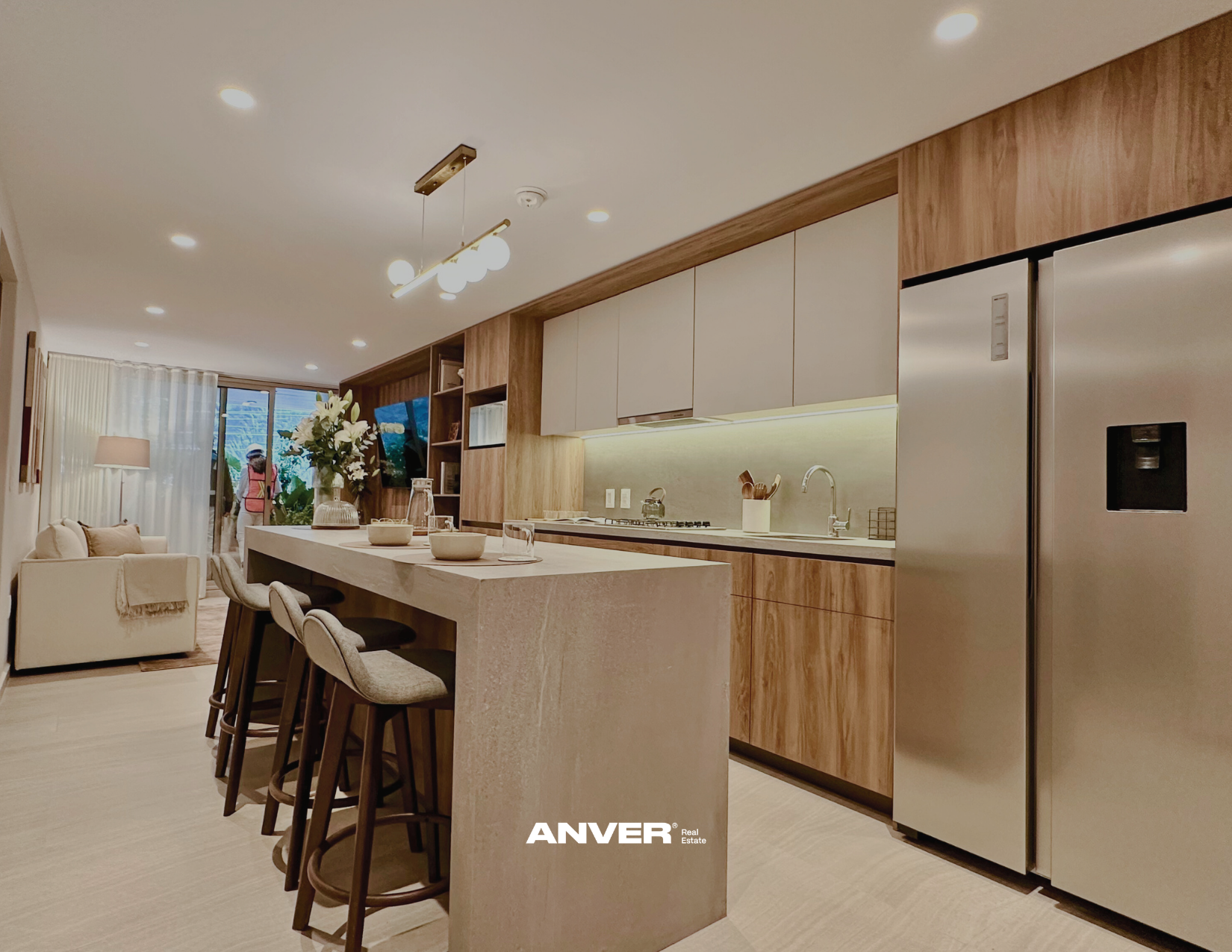 Cocina moderna con isla de piedra, gabinetes de madera y electrodomésticos de acero inoxidable en un espacio abierto que se conecta con la sala de estar, con una persona en el fondo.