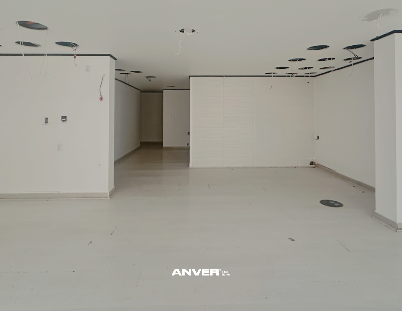 Interior de un espacio en construcción con paredes blancas, techo y piso claros, y orificios en el techo para luminarias.