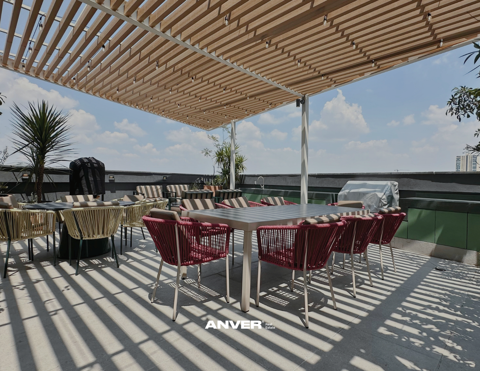 Terraza con mesas y sillas al aire libre bajo una pérgola de madera y luz solar, decorada con plantas y vista hacia el cielo y edificios en la distancia.