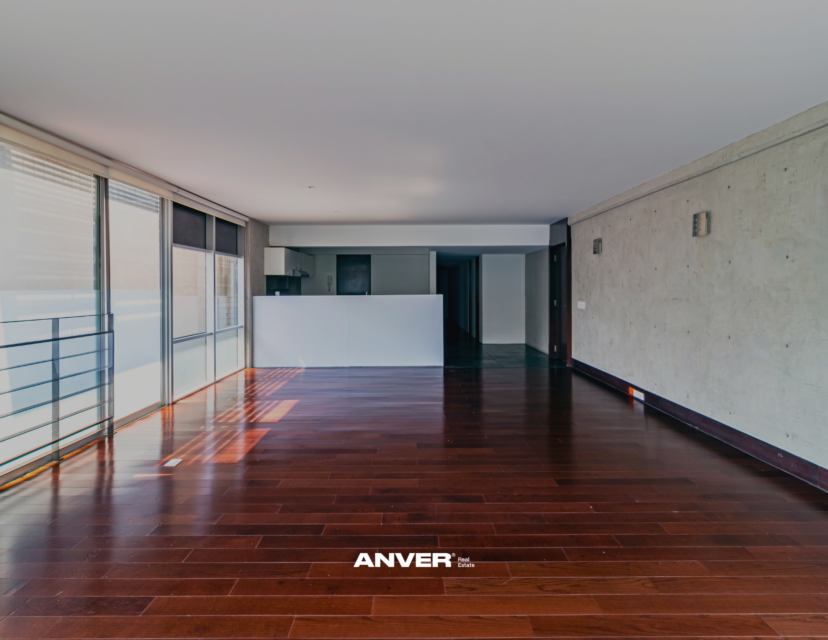 Espaciosa sala de estar con pisos de madera oscura, paredes de concreto y ventanales grandes con persianas. Cocina en el fondo y espacio vacío en el medio.