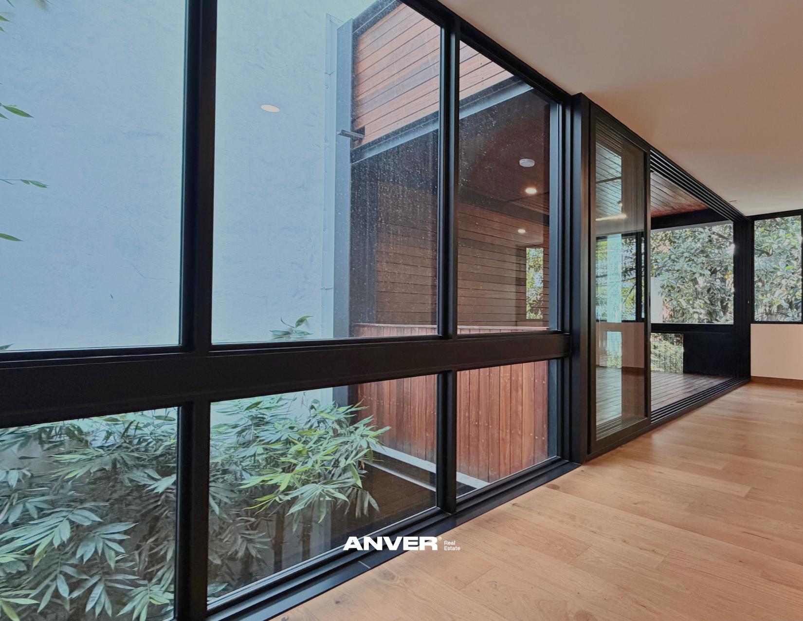 Vista interior de una sala con ventanas de vidrio negro que muestran un patio con vegetación y paredes de madera. Piso de madera clara y paredes blancas.