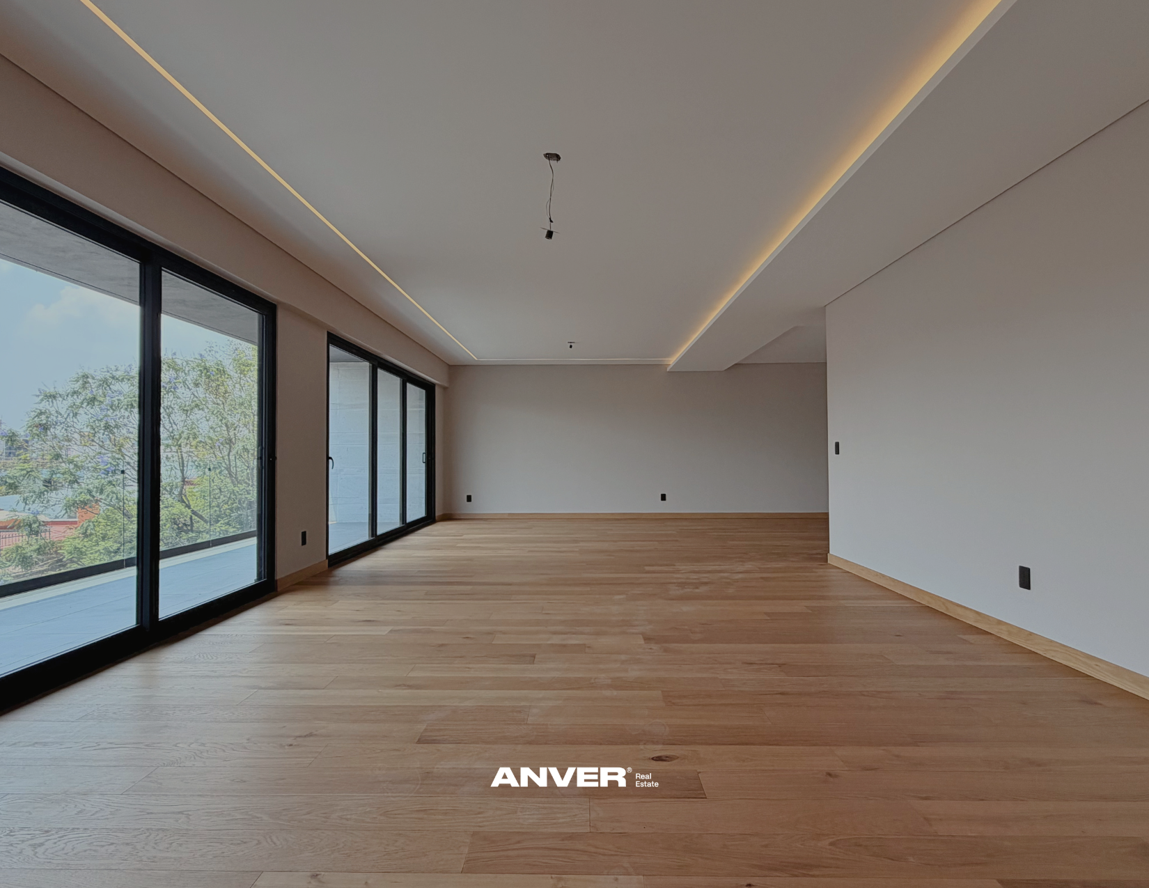 Espaciosa sala vacía con pisos de madera, paredes blancas y grandes ventanas correderas que dan a un balcón con vista a árboles y cielo despejado. El techo tiene iluminación empotrada y cableado sin lámpara instalada.