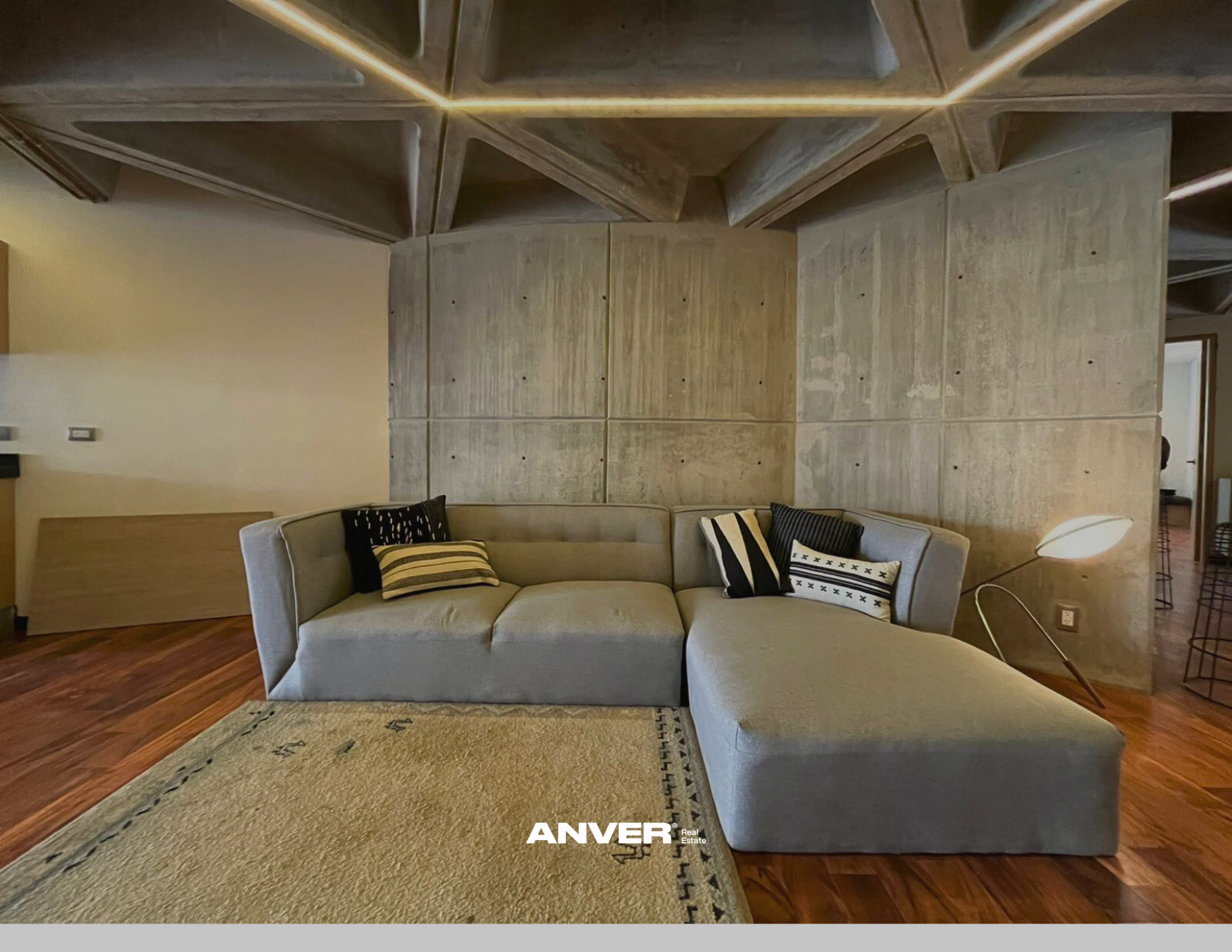 Sala de estar con sillón gris claro, cojines decorativos en tonos oscuros y rayados, una lámpara de lectura moderna, paredes de cemento y piso de madera.