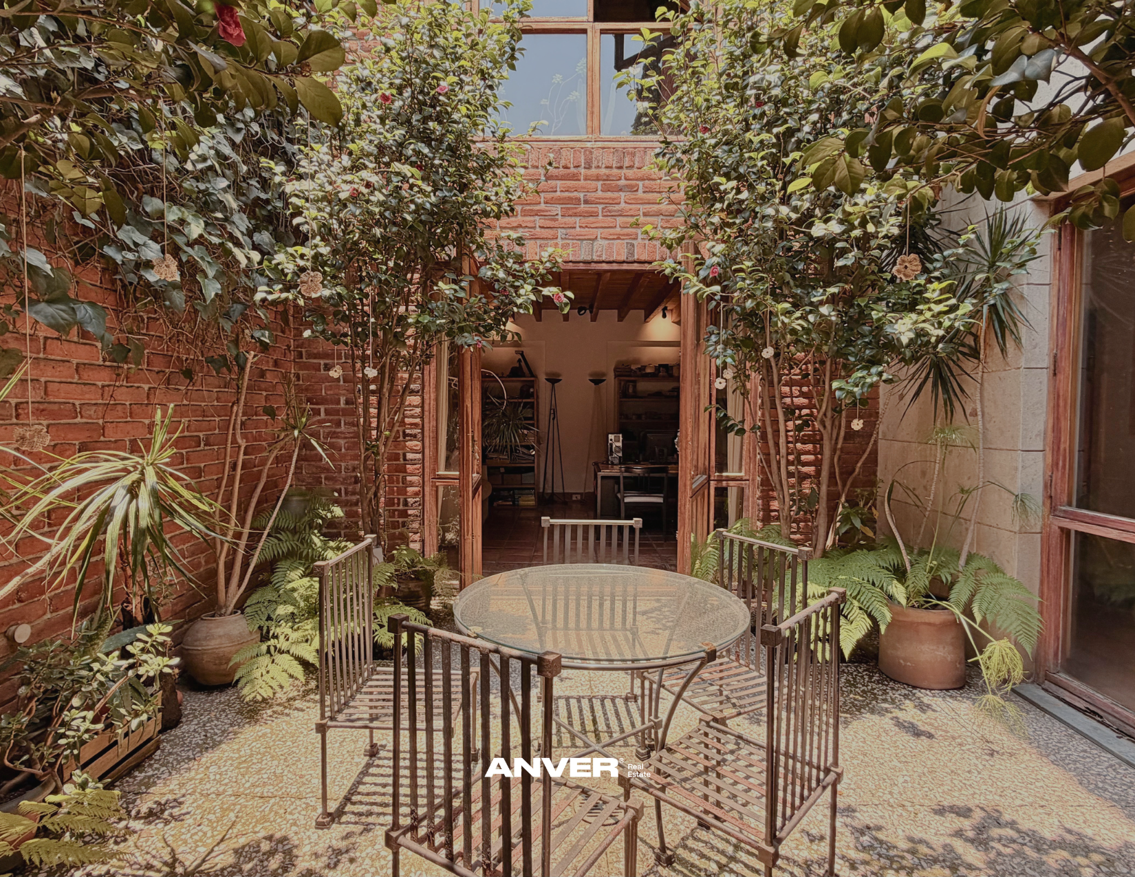 Patio con mesas de cristal, rodeado de plantas y paredes de ladrillo, con una puerta doble de madera que da a un interior