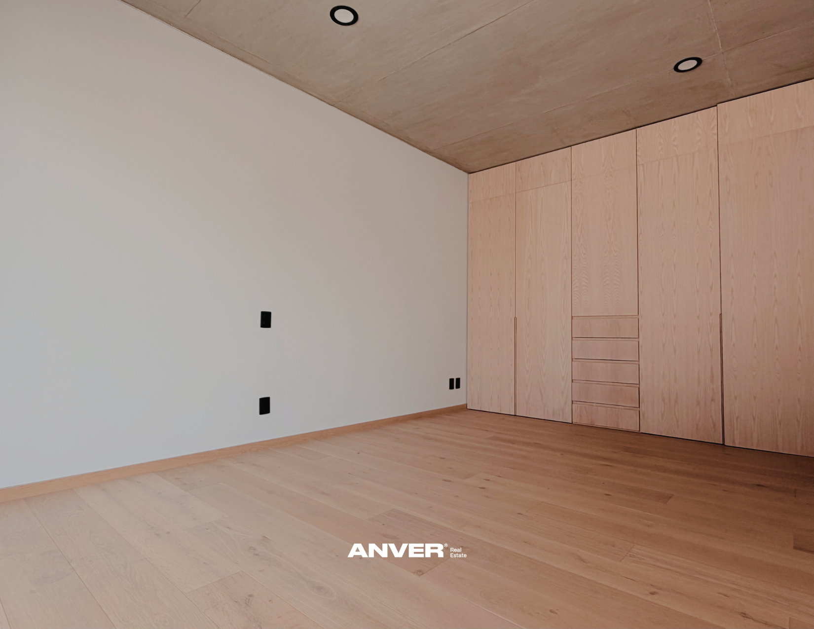 Habitación vacía con pisos de madera clara, paredes blancas y un armario de madera en una esquina. Cuatro enchufes y un interruptor visibles en la pared.