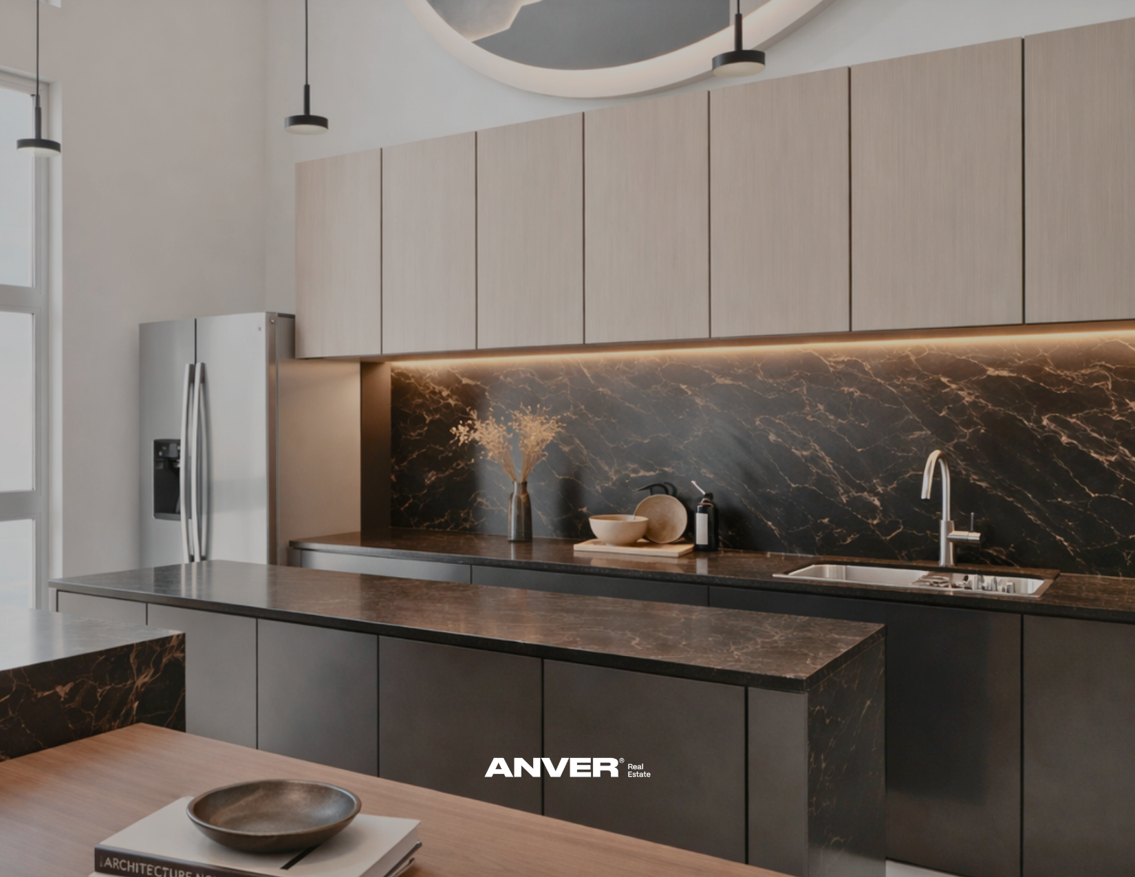 Cocina moderna con gabinetes de color claro, encimera de mármol oscuro con vetas doradas, azulejo de mármol oscuro en la pared, fregadero con grifo, y objetos decorativos como un jarrón con ramas secas, cuencos y un dispensador de jabón. Iluminación empotrada y lámparas colgantes en el techo.