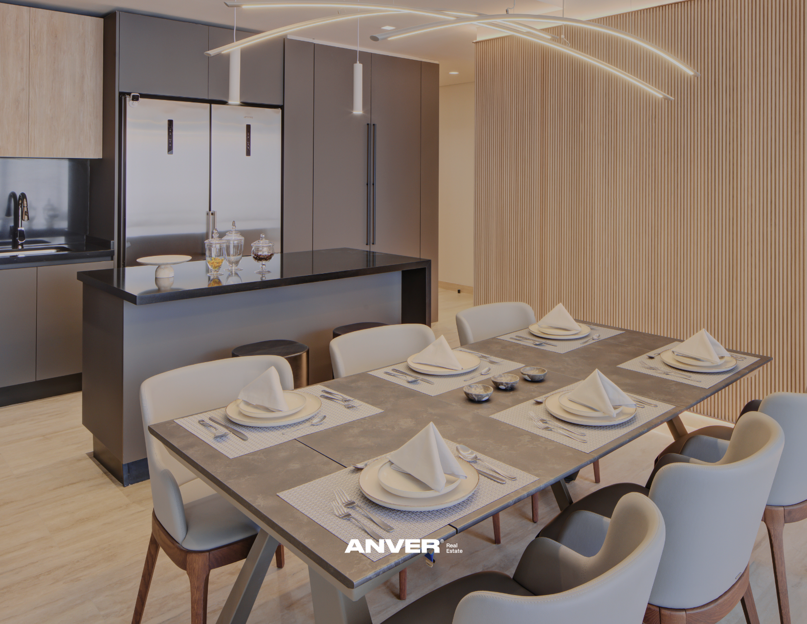Sala de comedor moderna con mesa que tiene seis lugares, platos blancos con servilletas dobladas, utensilios y pequeños recipientes decorativos. En el fondo, una cocina con electrodomésticos de acero inoxidable, armarios en tonos grises y madera clara, y un pared de paneles de madera vertical. La iluminación es elegante y lineal.