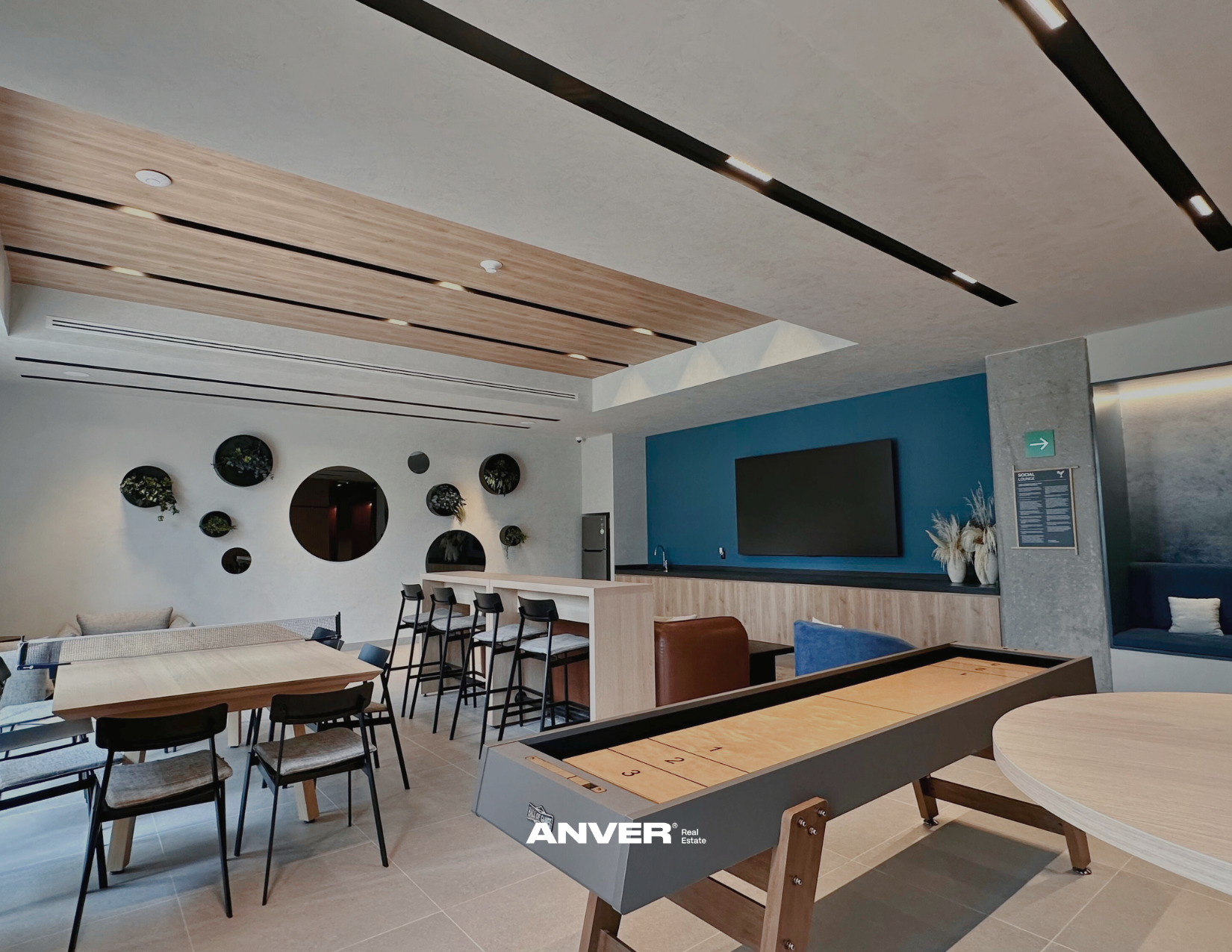 Sala de descanso con mesas y sillas, paredes decoradas con círculos y plantas, pantalla de televisión en la pared, zona de bar con lavabo y plantas decorativas, y juego de aire hockey en primer plano.