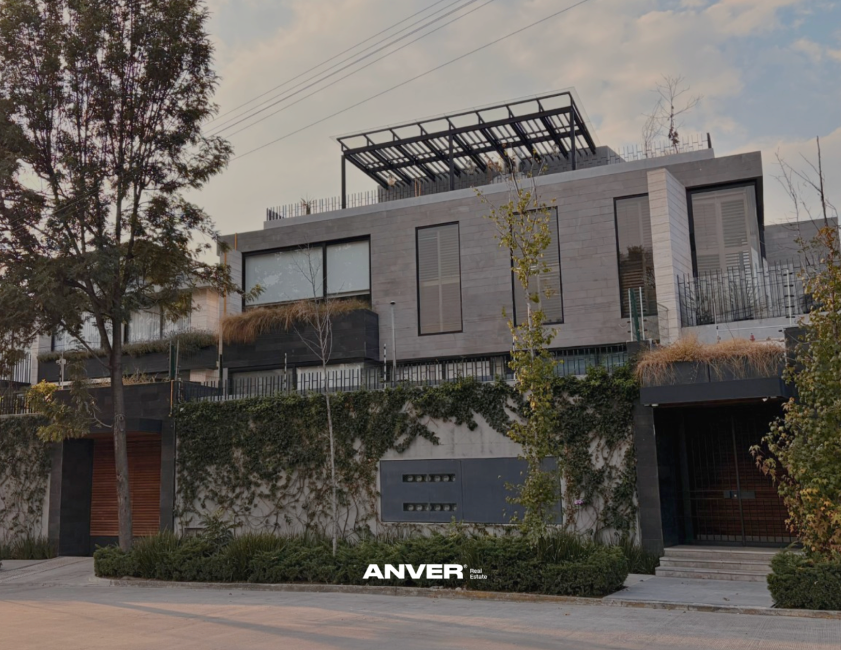 Edificio moderno de varias plantas con fachada de piedra y ventanas grandes, rodeado de árboles y vegetación, con una entrada de escalones y un portón de acceso, y un cielo parcialmente nublado.