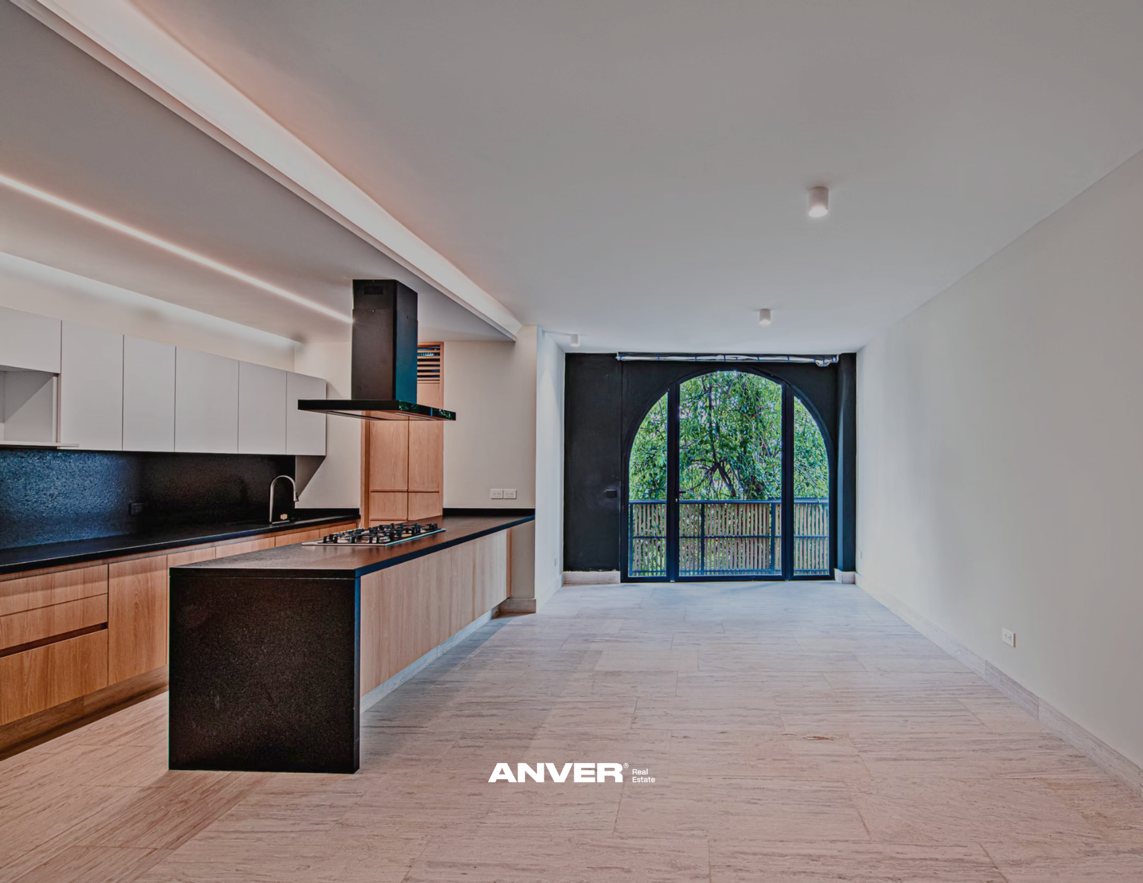 Cocina moderna con gabinetes de madera clara, encimera negra, estufa de gas y campana extractora negra, y gran puerta de arco negra que lleva a un balcón con vista a árboles verdes.
