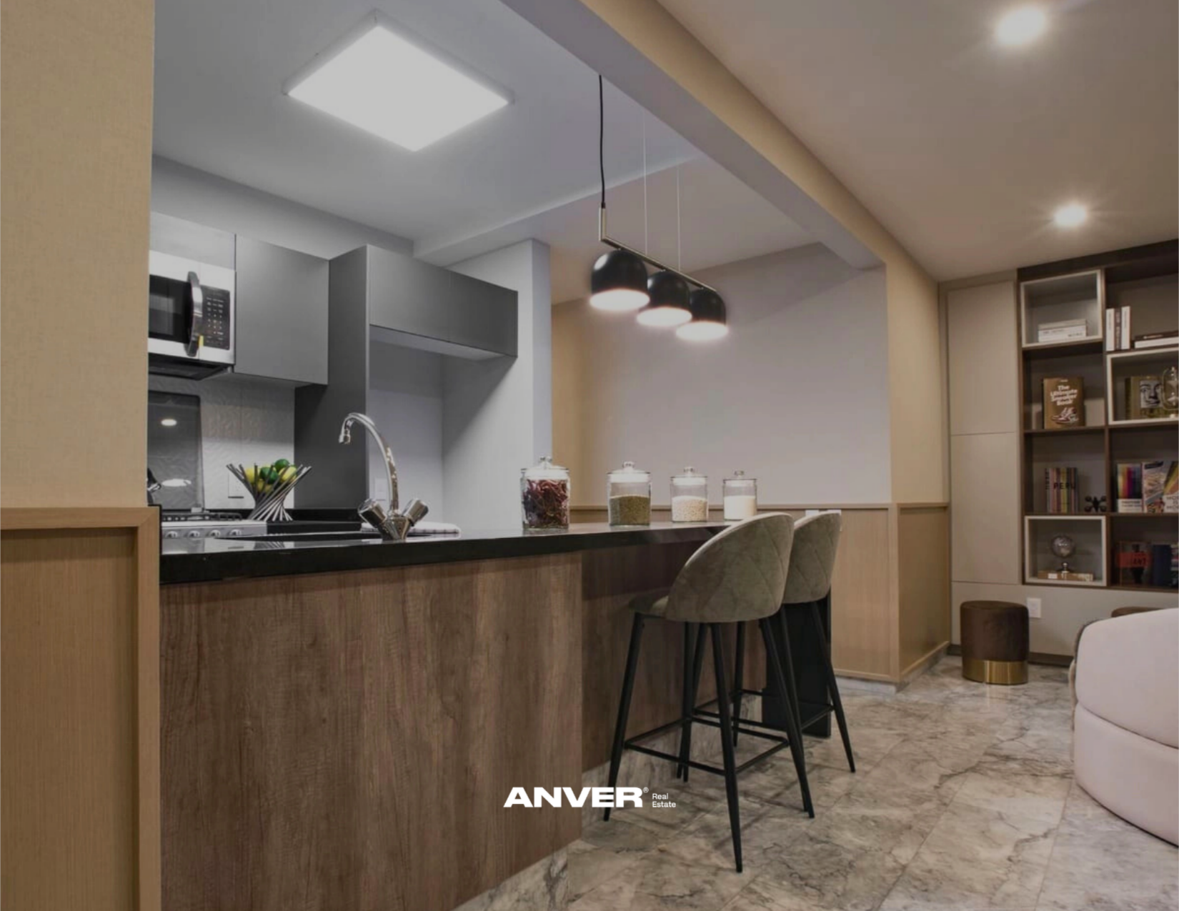 Cocina moderna con barra de desayuno, cinco frascos con ingredientes, y sala con biblioteca y sillón.