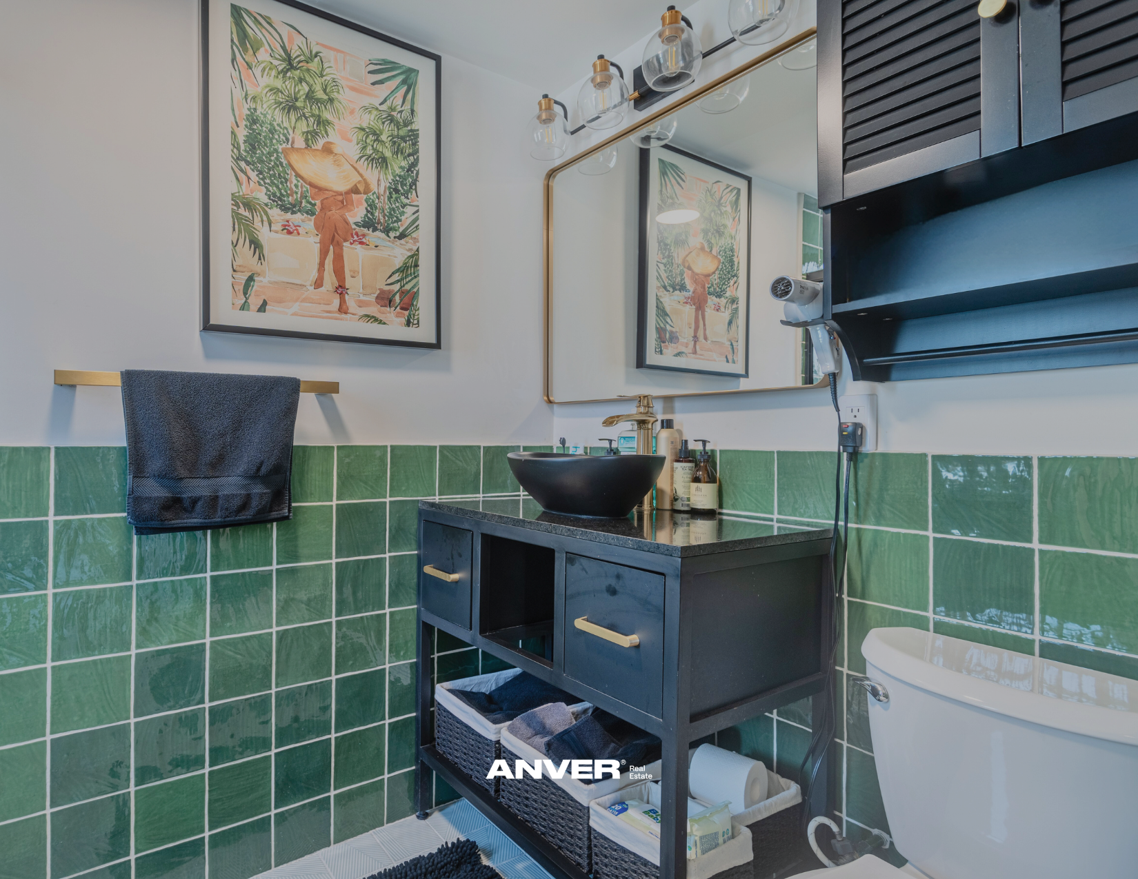 Un baño moderno con pared de azulejos verdes y un lavabo negro en una encimera negra. Encima hay ungüentos y un lavabo negro en la pared en ángulo con un espejo grande. Una toalla gris en un soporte dorado y una lámpara colgante de seis bombillas de vidrio y detalles dorados. Una ilustración de un hombre con sombrero en la pared, y una caja de almacenamiento debajo del lavabo con papel higiénico y productos de limpieza.