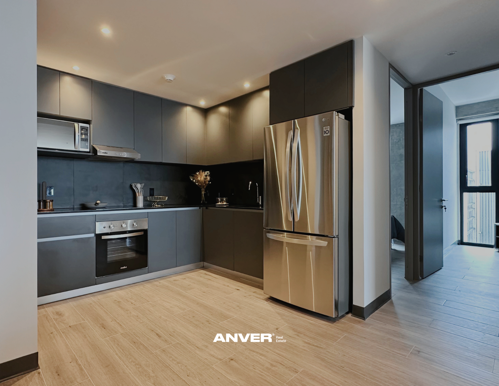 Cocina moderna con gabinetes en tonos grises, electrodomésticos de acero inoxidable, un horno, microondas y refrigerador. Piso de madera clara y área de entrada con puerta y ventana al fondo.