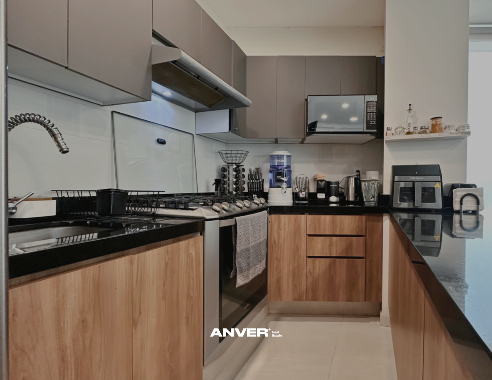 Cocina moderna con gabinetes de madera, encimera negra, electrodomésticos de acero inoxidable, y utensilios en la encimera.