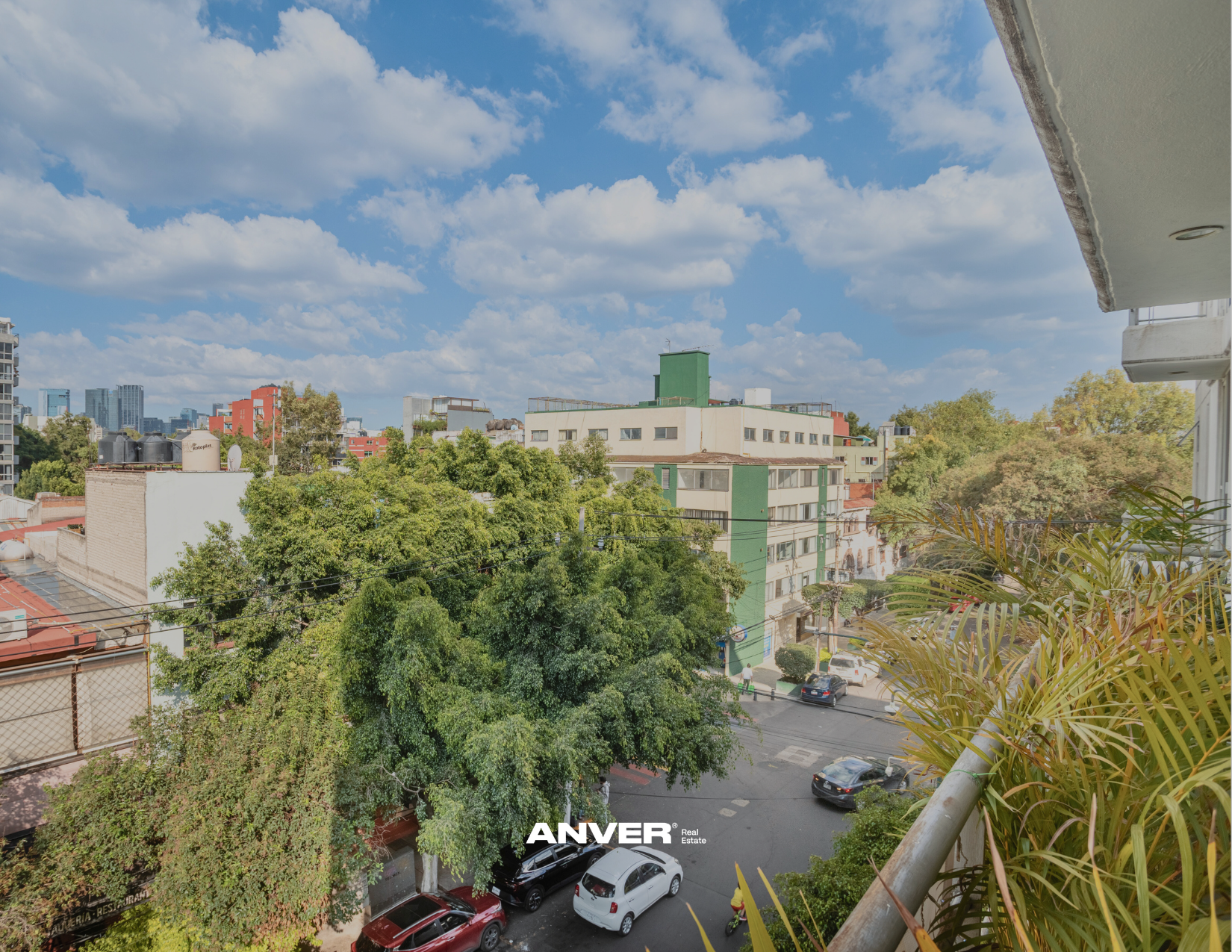 Vista de un barrio urbano con árboles, edificios y calles, vista desde un balcón con vegetación en primer plano y un cielo azul con nubes.
