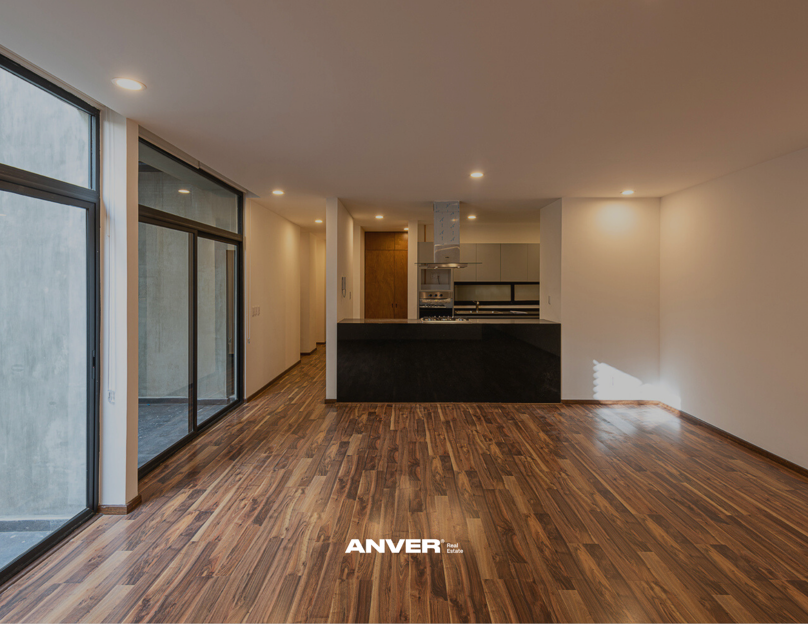 Interior de una casa moderna con piso de madera, paredes blancas, grandes ventanas de vidrio y una cocina en el fondo.