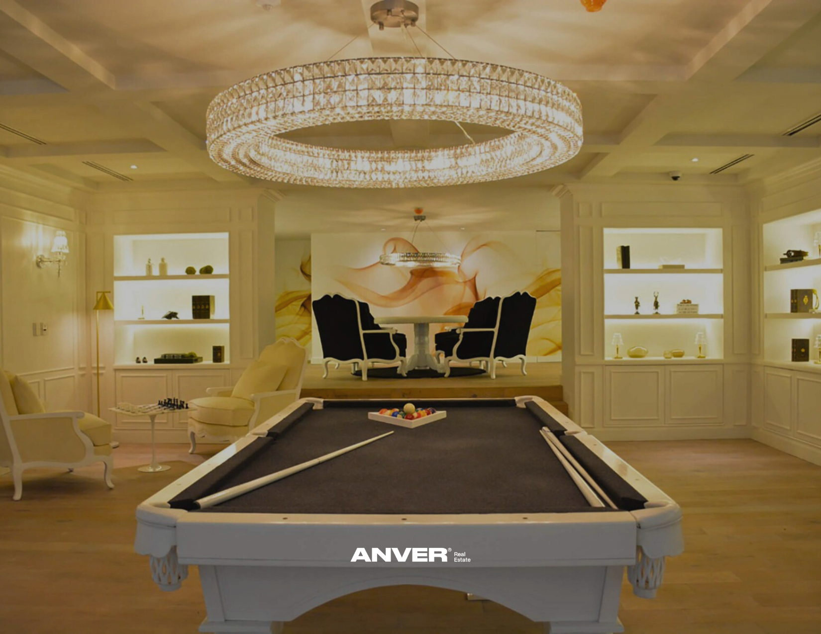 Sala de juegos con mesa de billar, sillones blancos, estanterías con decoraciones y una lámpara de araña grande en el techo.