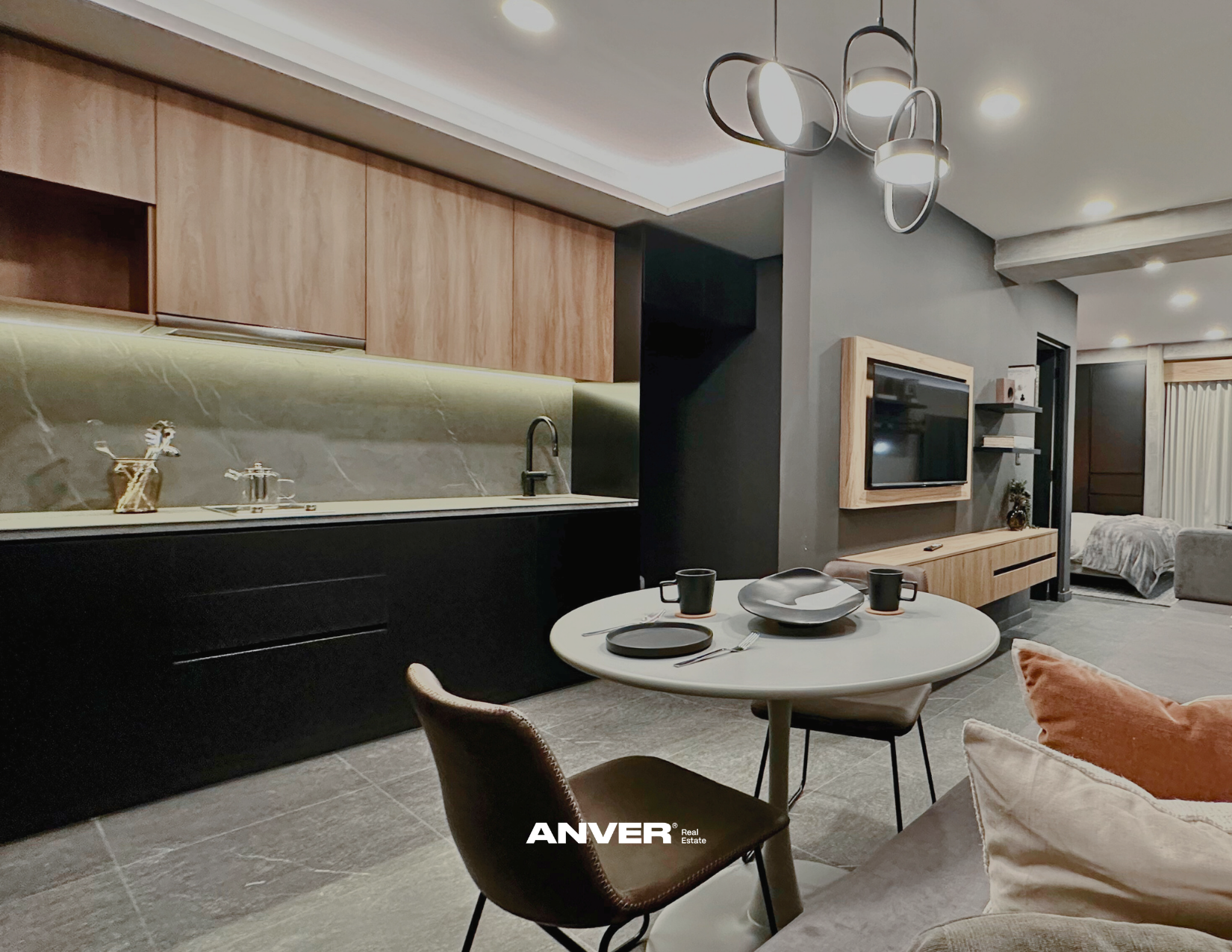 Vista de una sala moderna con cocina integrada, mesa redonda con utensilios y tazas, sillón con cojín, televisión en la pared y habitación con cama en el fondo.