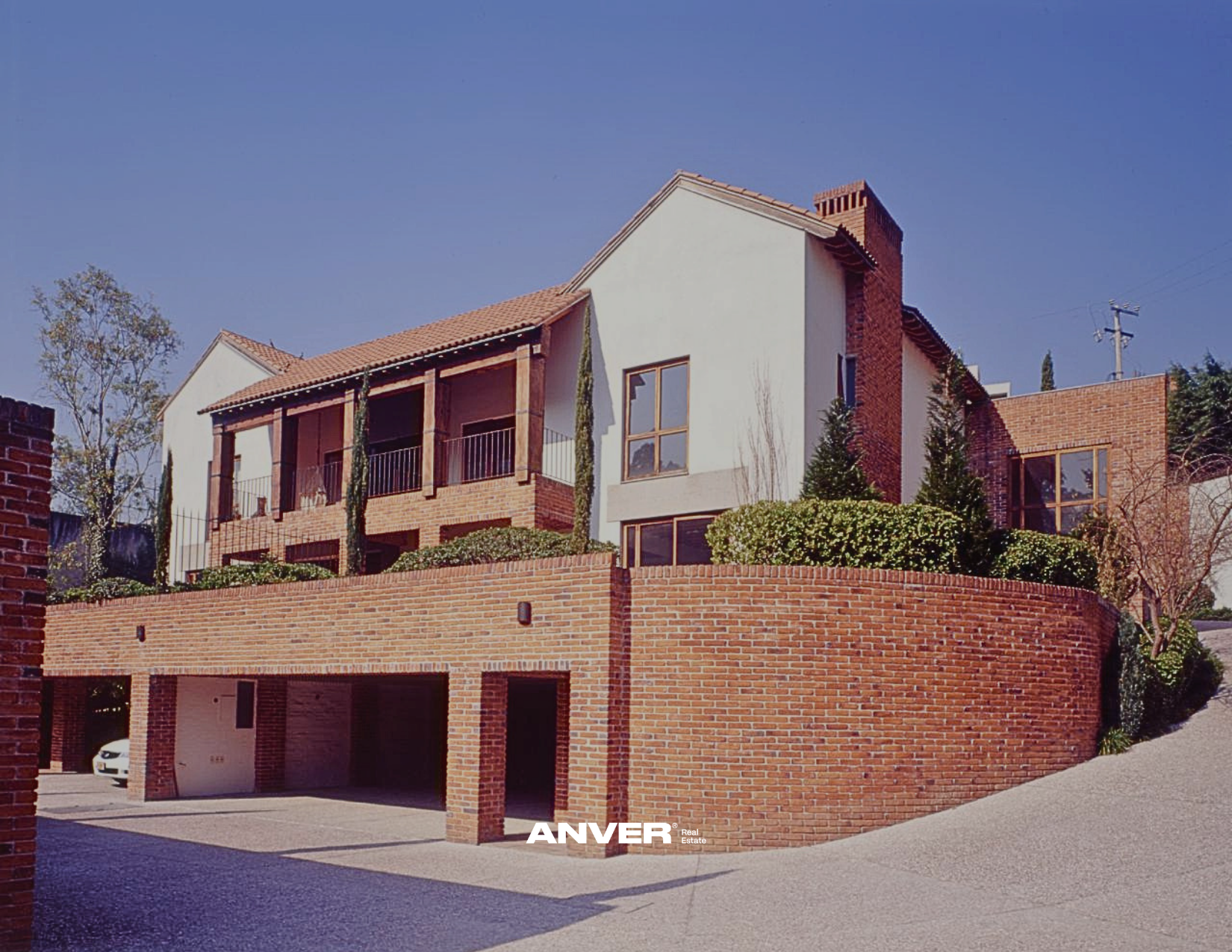 Casa moderna de varias plantas con fachada de ladrillo y paredes blancas, jardines con arbustos y árboles, y un garaje en la planta inferior, bajo un cielo despejado.