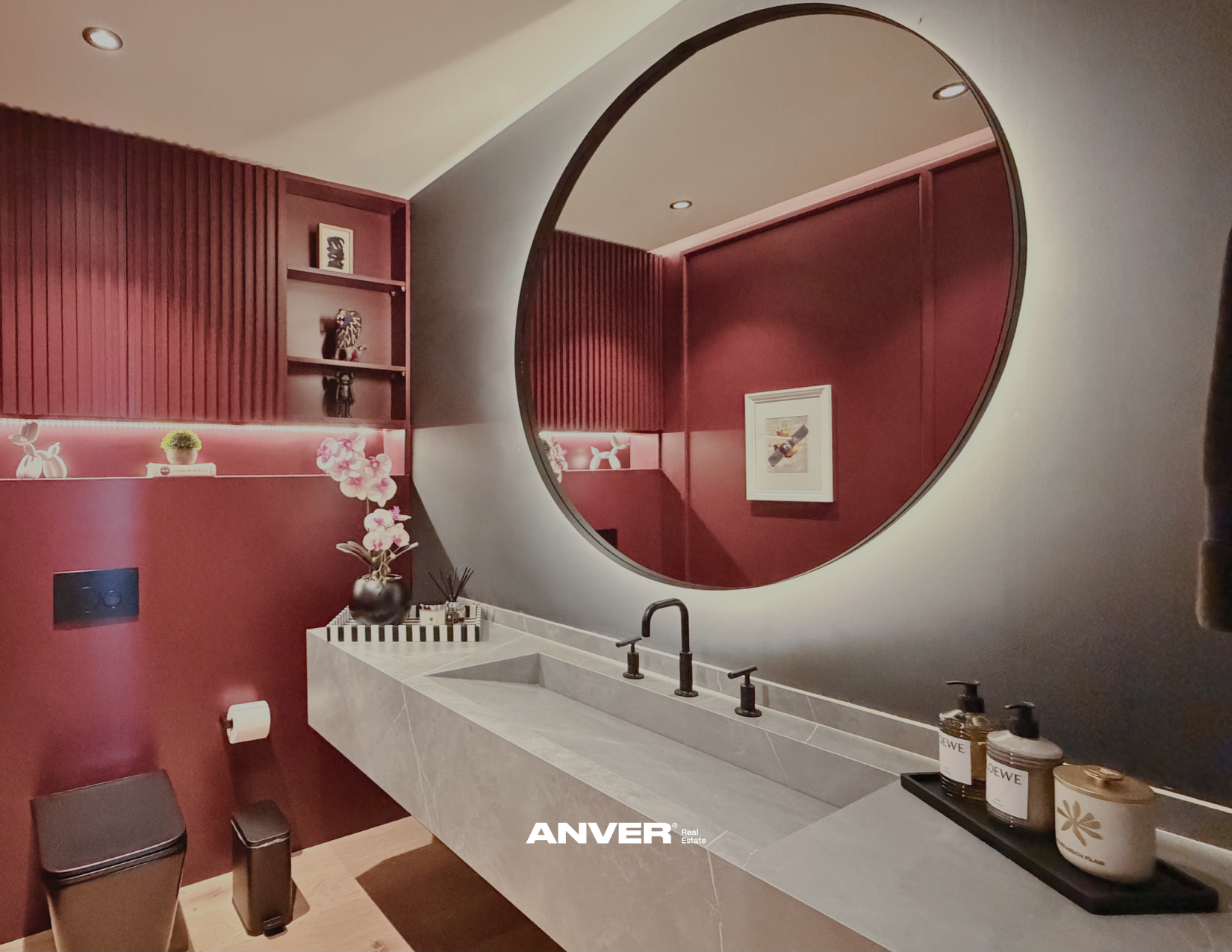 Espejo grande en forma circular en pared gris, lavabo de concreto con accesorios negros, paredes rojas con estanterías decorativas y planta, en un baño moderno y elegante.