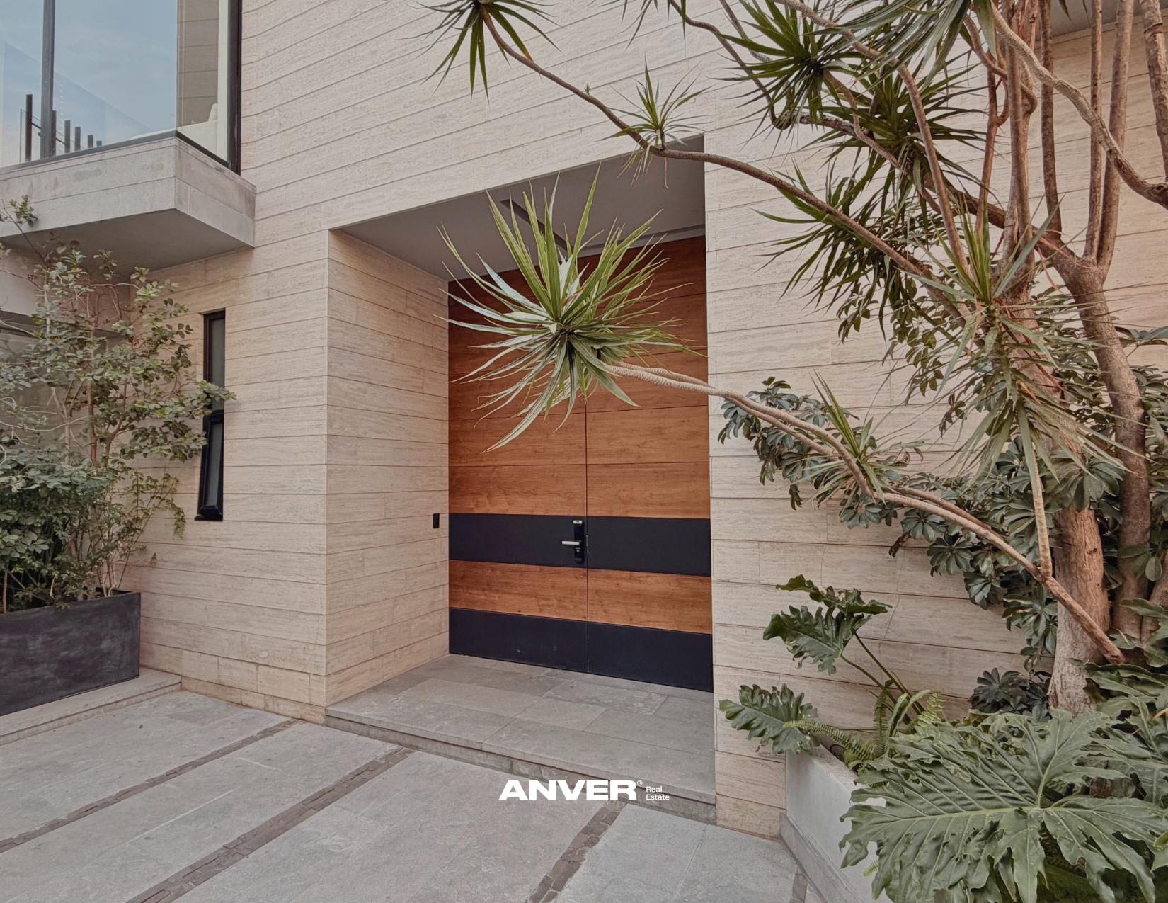 Entrada moderna a una vivienda con puerta de madera y detalles en negro, rodeada de plantas verdes en macetas y en el suelo, con paredes de piedra clara.