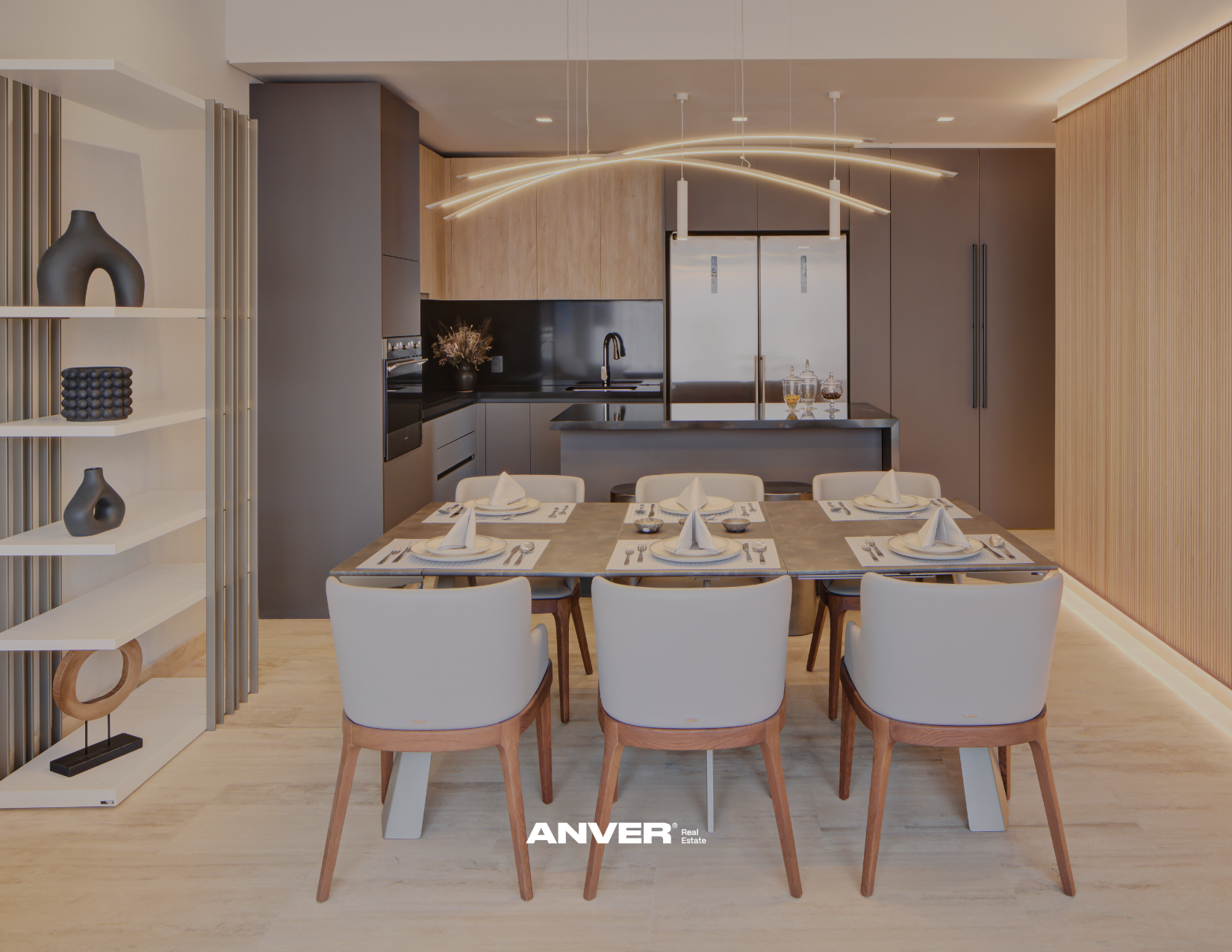 Interior de una sala comedor moderna con mesa puesta para seis personas, en el fondo una cocina con gabinetes en tonos madera y gris, y lámparas decorativas en el techo. La imagen tiene el logo de ANVER Real Estate en la parte inferior.