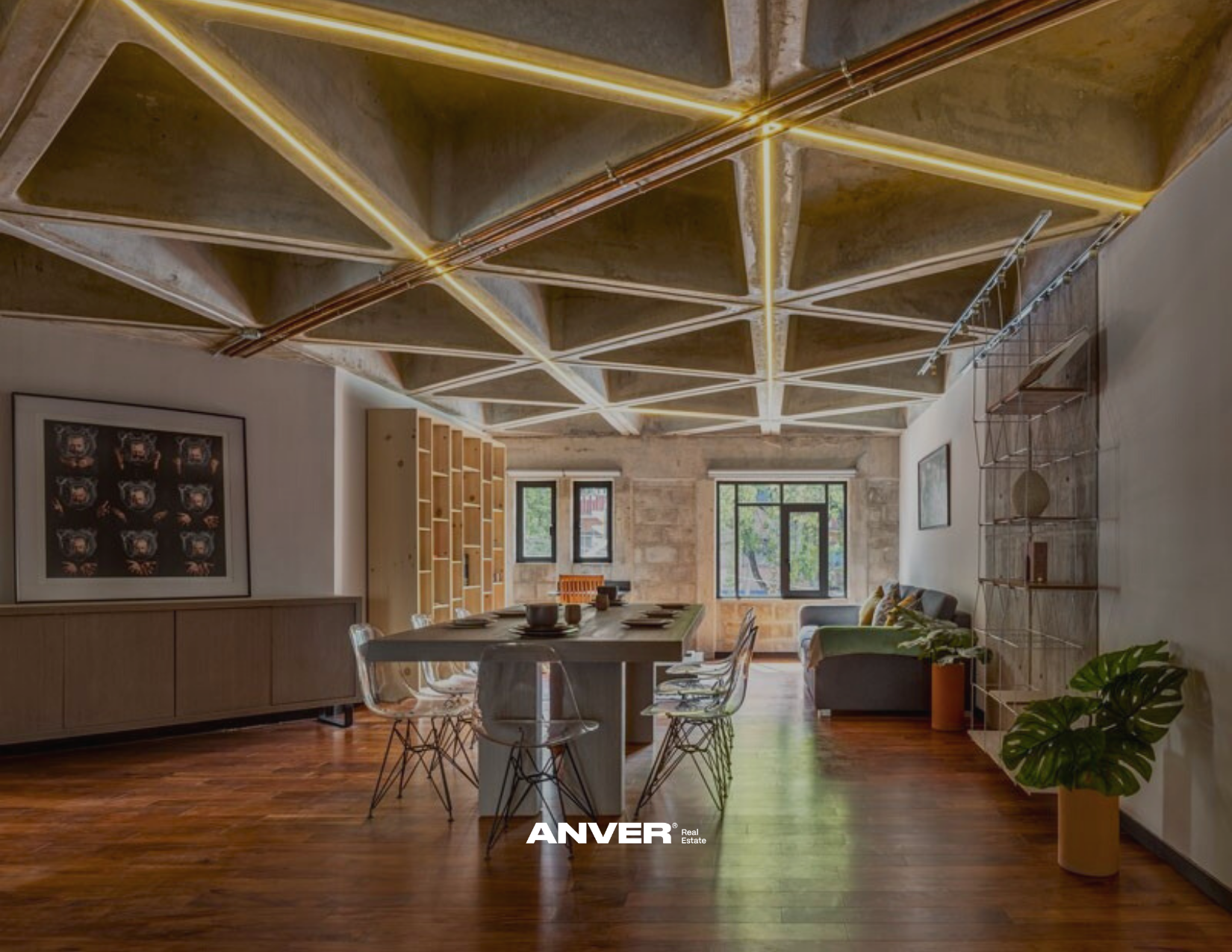 Interior de un espacio moderno con mesa de comedor rectangular, sillas transparentes, sofá gris, estantería de madera y paredes de piedra y blanco, ventanas grandes, con plantas y arte en las paredes.
