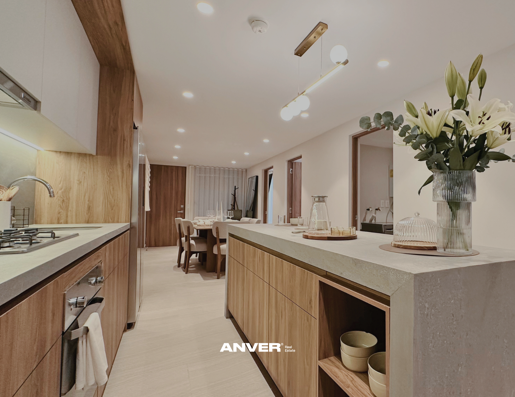 Interiores de una cocina moderna con madera clara, encimera de piedra, una flor blanca en un jarrón y vista a un comedor con sillas de madera y paredes blancas.