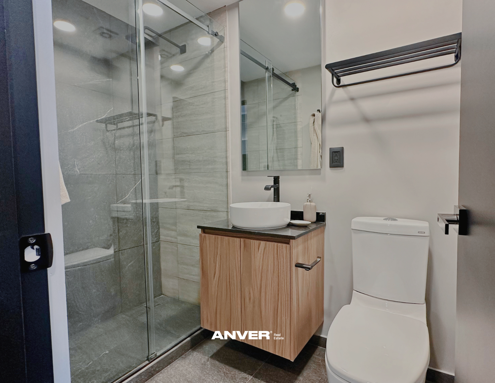 Baño con ducha con puertas de vidrio, lavabo sobre mueble de madera, espejo y retrete en un diseño moderno y minimalista.