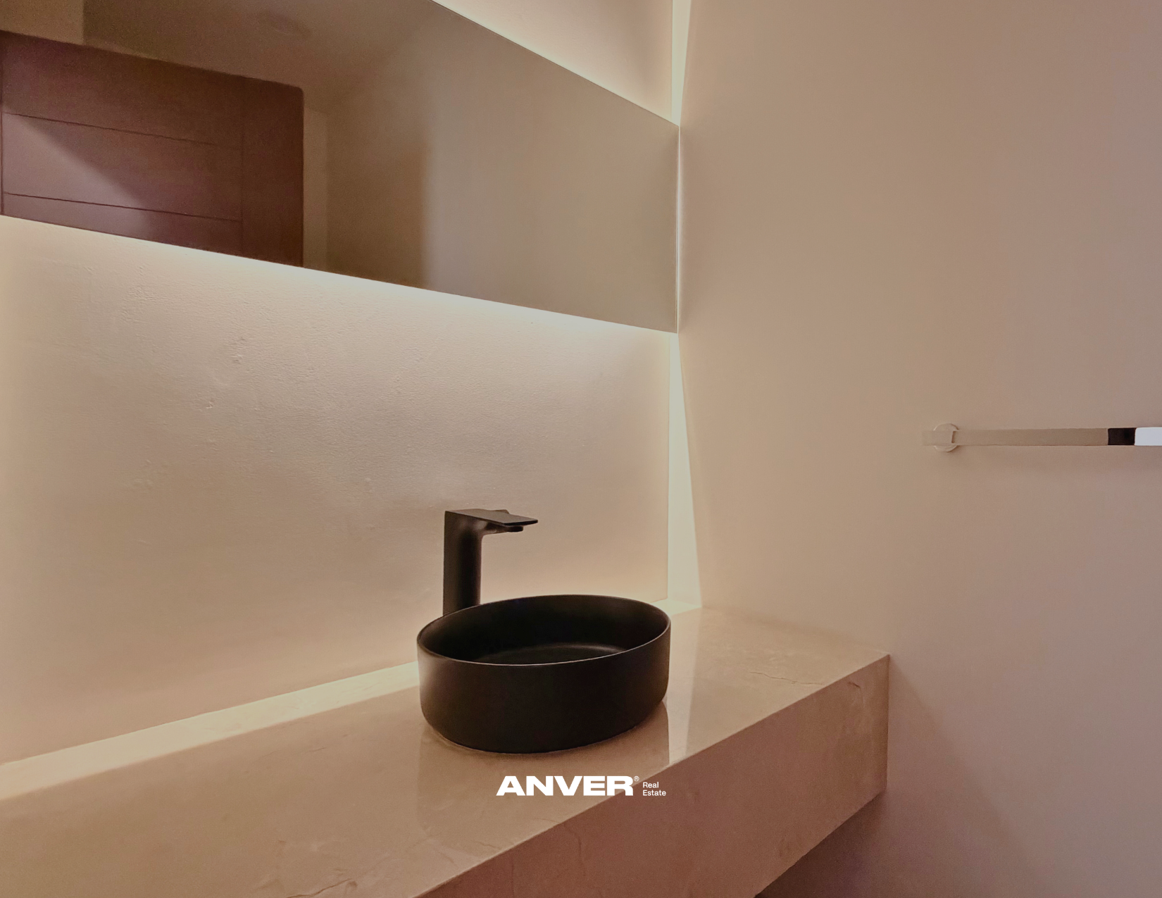 Lavabo negro sobre encimera de piedra en un baño moderno con paredes blancas y lámparas de luz indirecta.