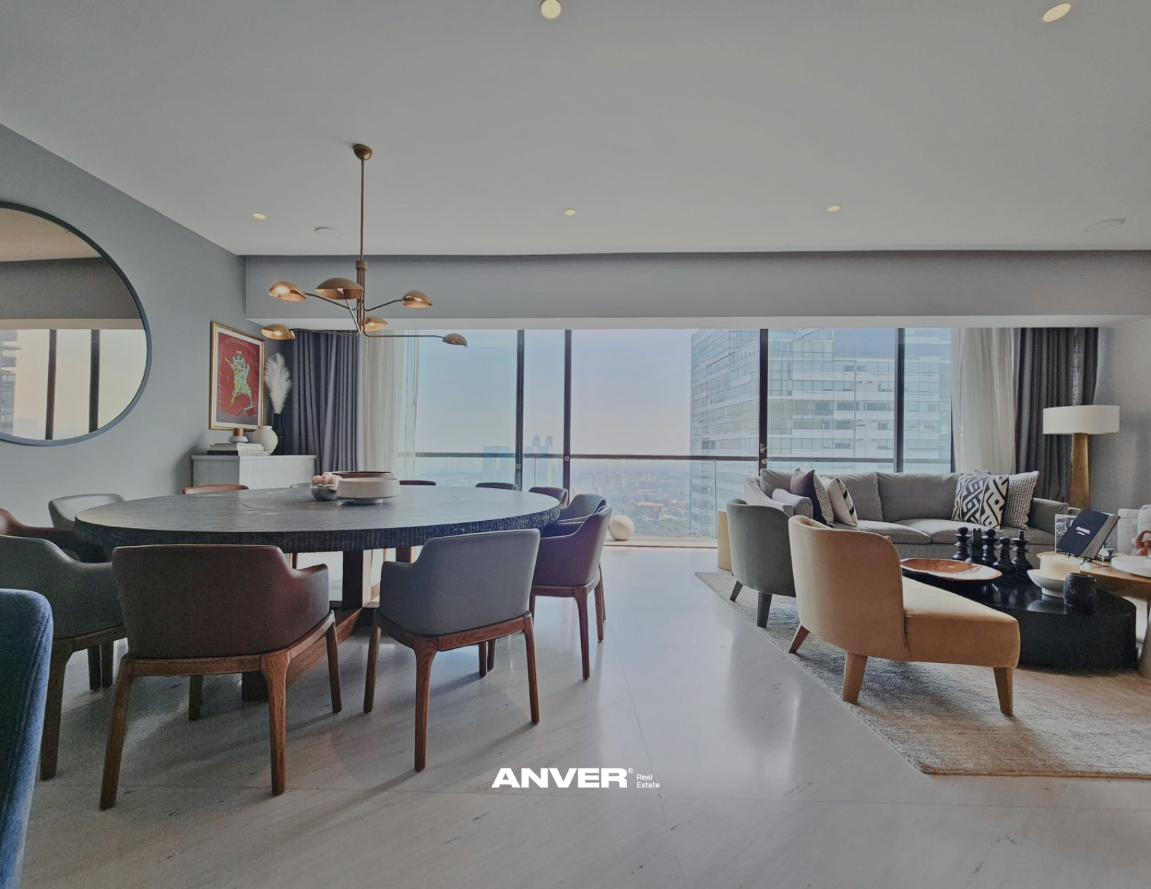 Interior de una sala moderna con sillones, una mesa redonda y una sala de estar con sofá y mesas, con ventanas grandes y vista a edificios urbanos.