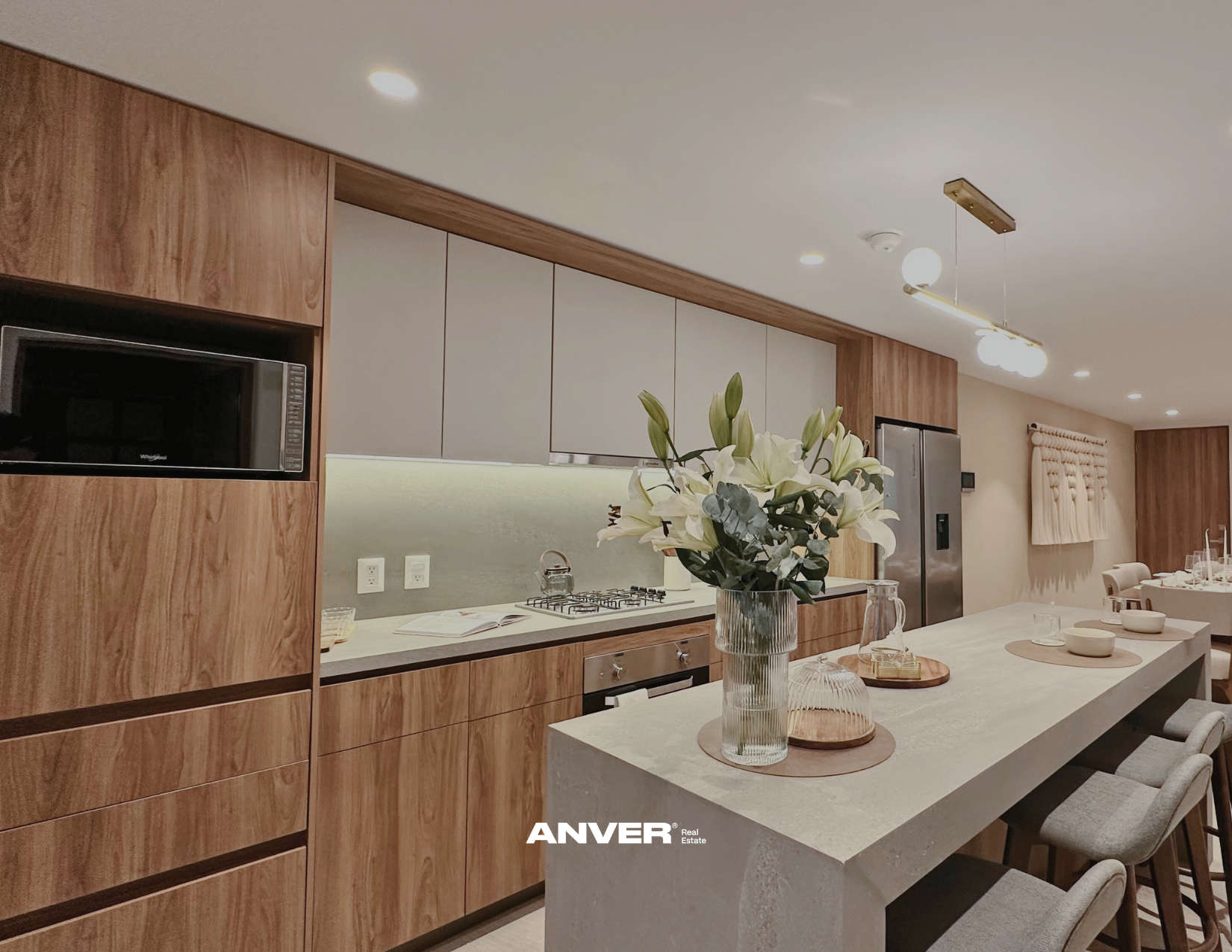 Cocina moderna con isla central, encimeras de piedra, electrodomésticos de acero inoxidable, y decoración con flores blancas en un jarrón de cristal.