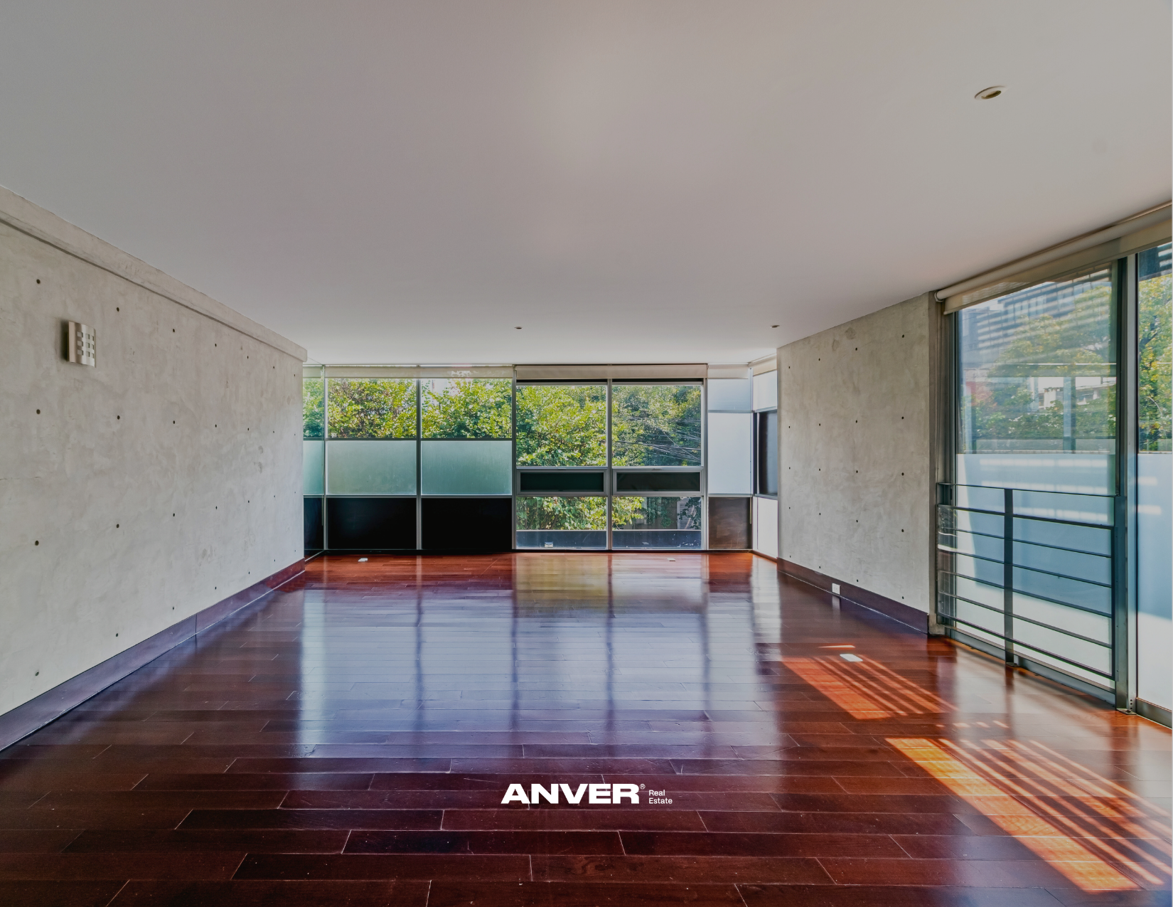 Sala vacía con pisos de madera oscura y paredes de concreto, ventanas grandes con vista a árboles, y logo de ANVER en la parte inferior.