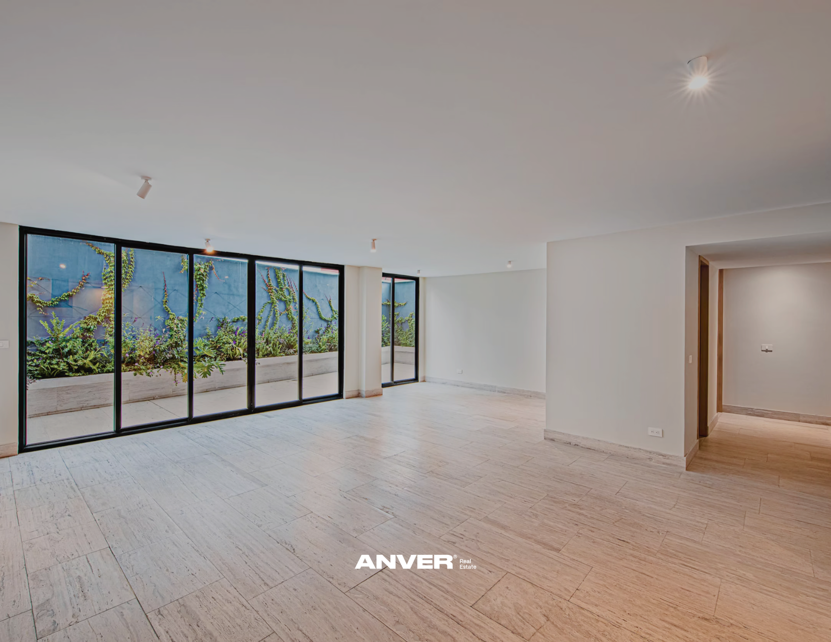 Espacio interior vacío con paredes blancas, piso de baldosas claras, grandes ventanales con vista a un patio con plantas y arbustos, entrada a otra habitación a la derecha, luces en el techo.