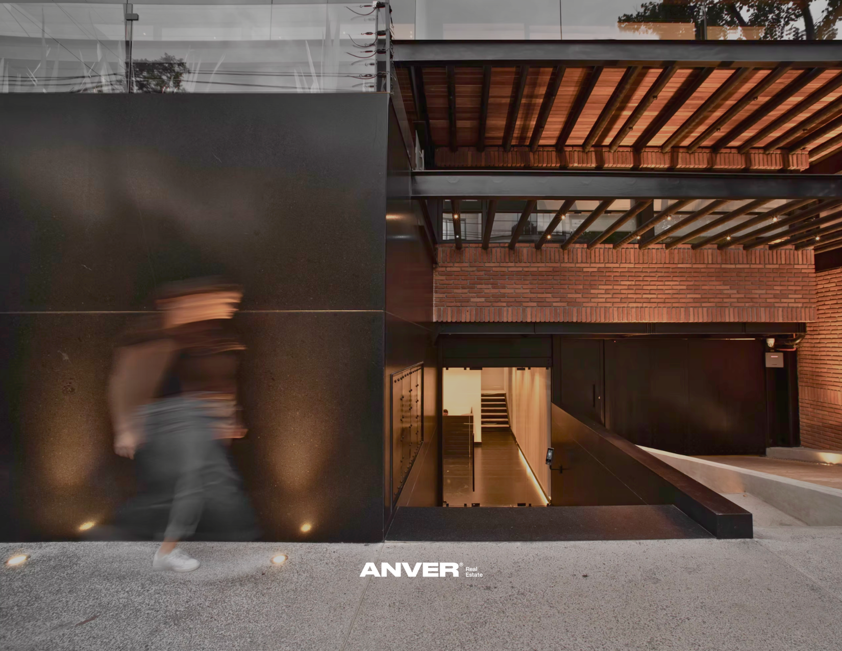 Vista de un edificio moderno con fachadas de ladrillo y ventanas de cristal, entradas y escaleras internas visibles, y un joven caminando rápidamente en primer plano, con iluminación artificial en el interior.