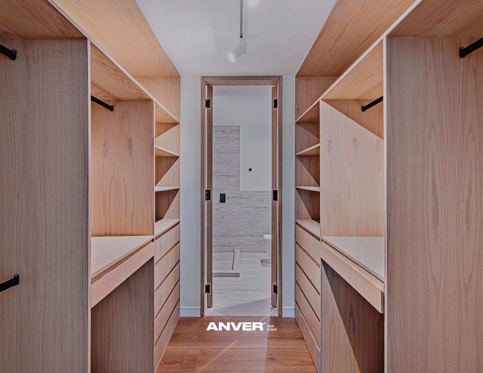 Vestidor de madera con estantes y cajones a ambos lados, puertas de madera al fondo que llevan a un baño con paredes de piedra y un inodoro.