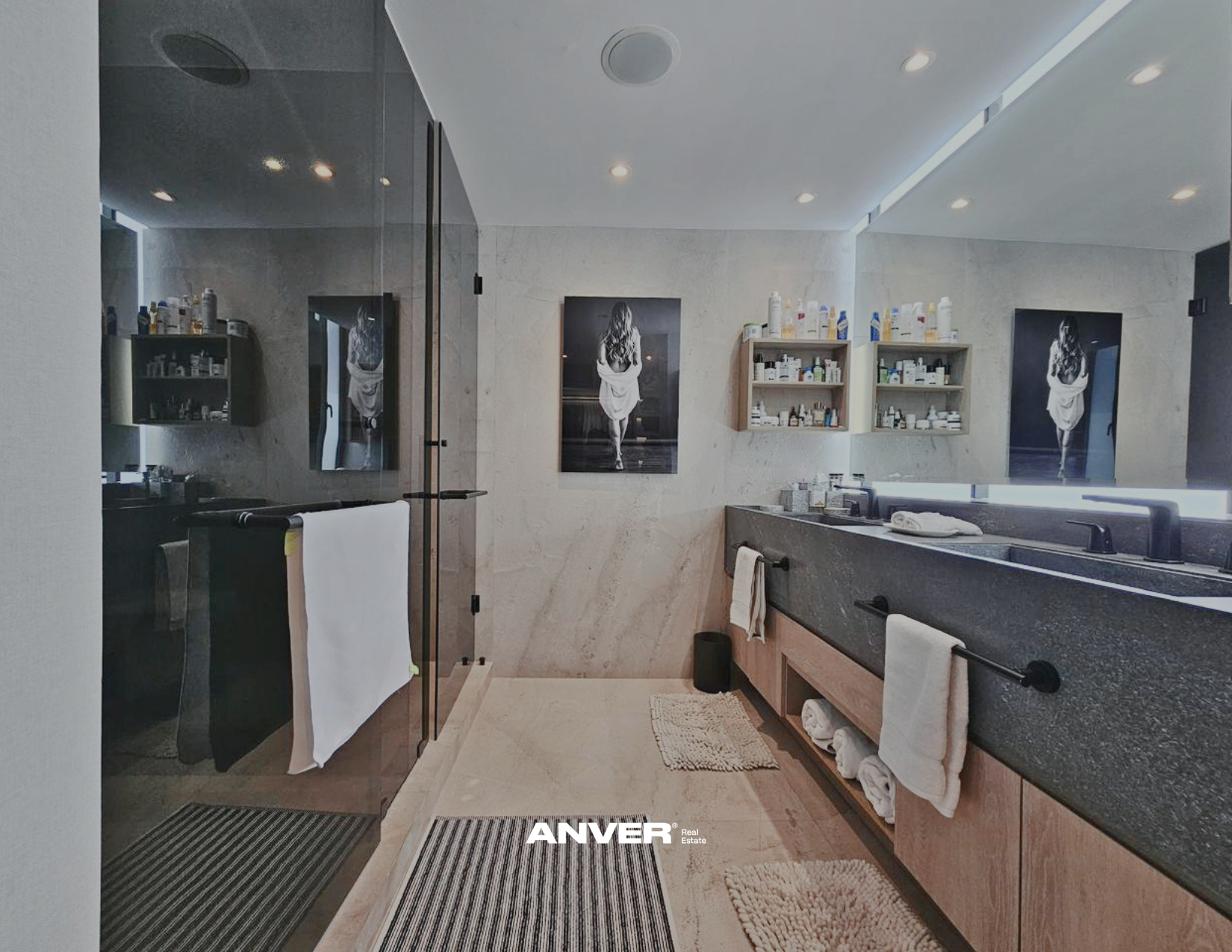 Baño moderno con paredes de piedra, espejo grande, armarios con productos de higiene, toallas colgadas, fotografía en blanco y negro de una mujer caminando, y un espejo con iluminación LED.