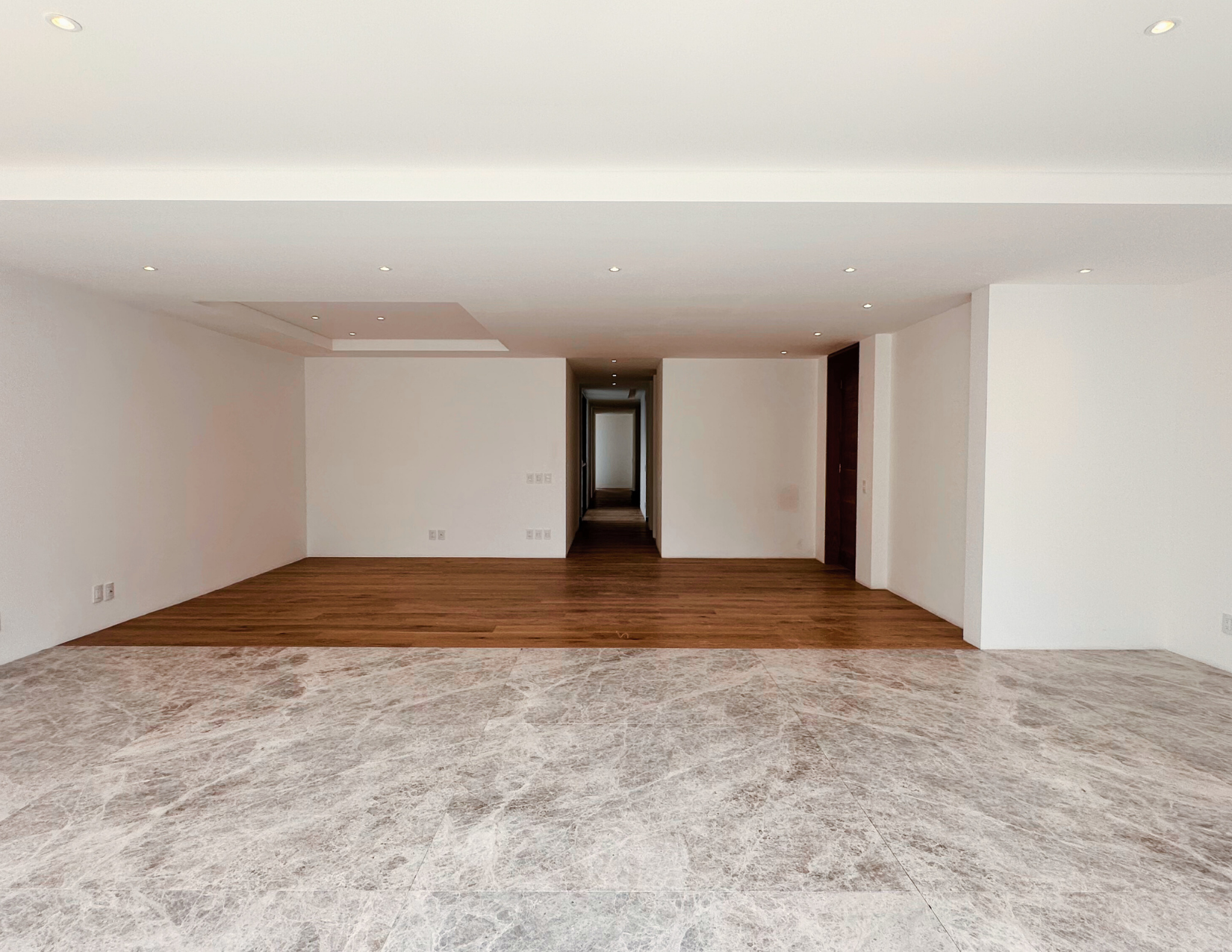 Habitación vacía con paredes blancas, suelo de madera y una alfombra en primer plano, con puertas al fondo y luces en el techo.