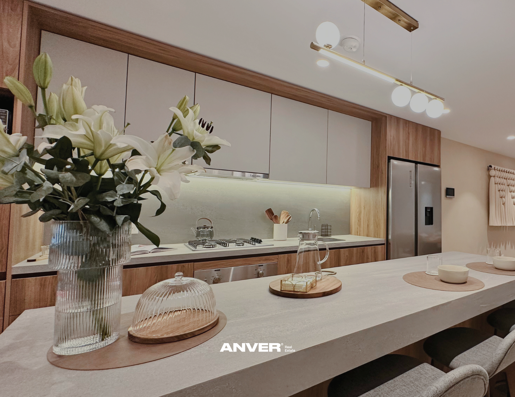Vista de una cocina moderna con muebles de madera clara, con flores blancas en un florero de vidrio en la esquina izquierda, y en la mesa hay bandejas redondas de madera con platos, tazas y una jarra, con iluminación moderna en el techo.
